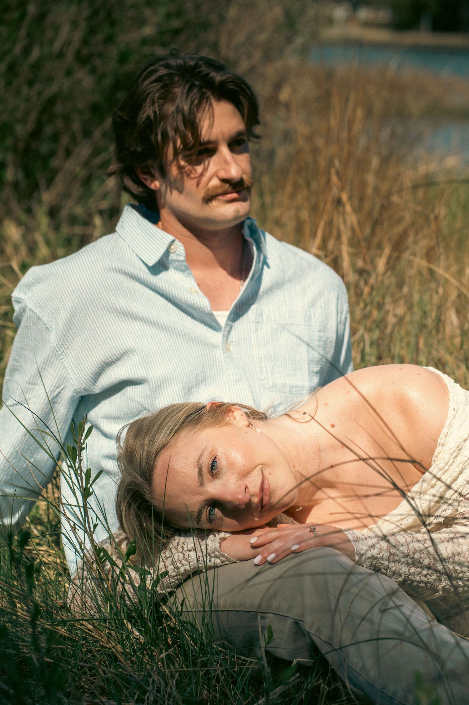 A couple relaxes in tall grass near water, with the woman resting her head on the mans lap.