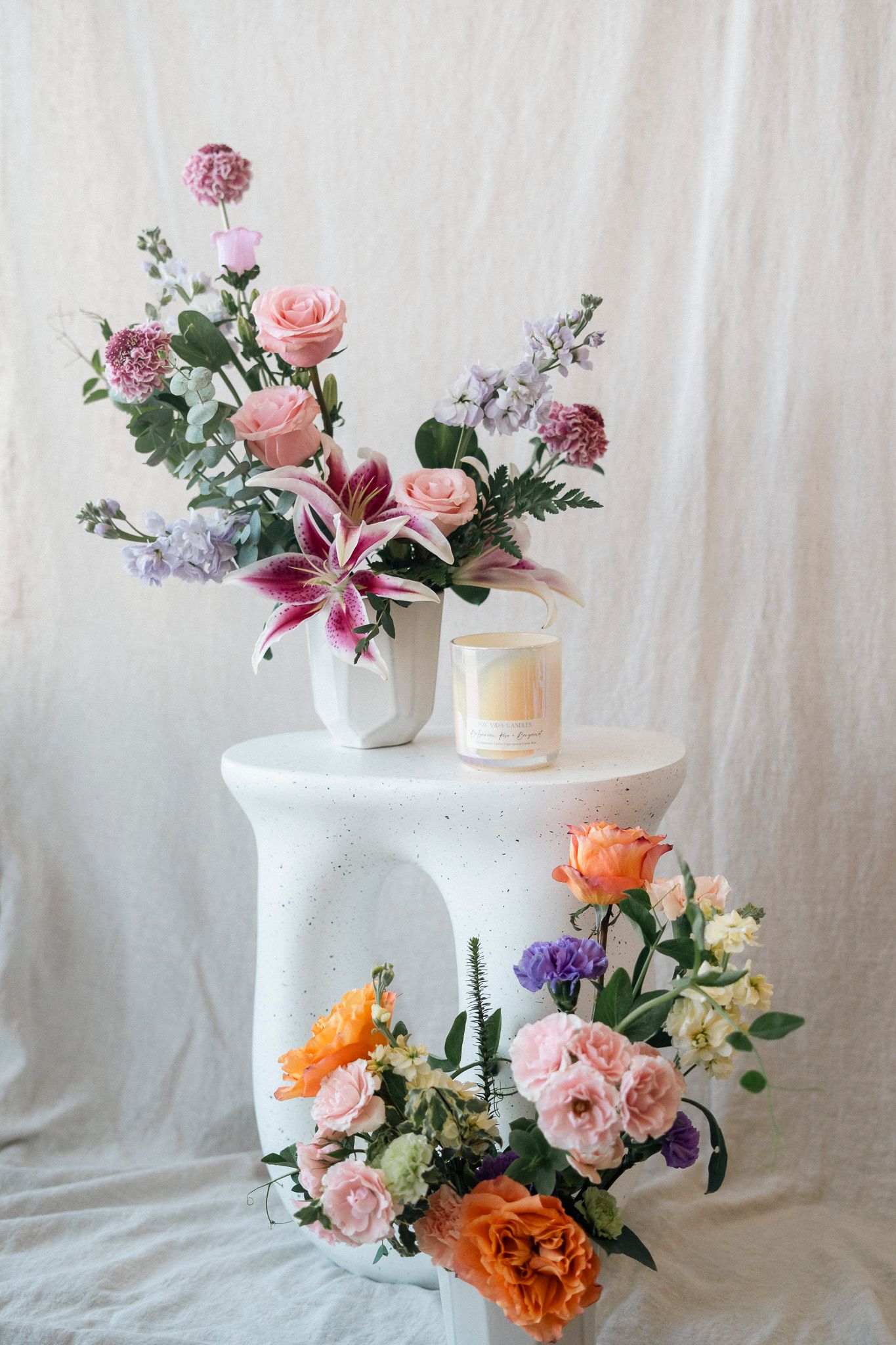 Elegant floral arrangement with pink roses, lilies, and purple flowers in a white vase beside a candle on a pedestal.