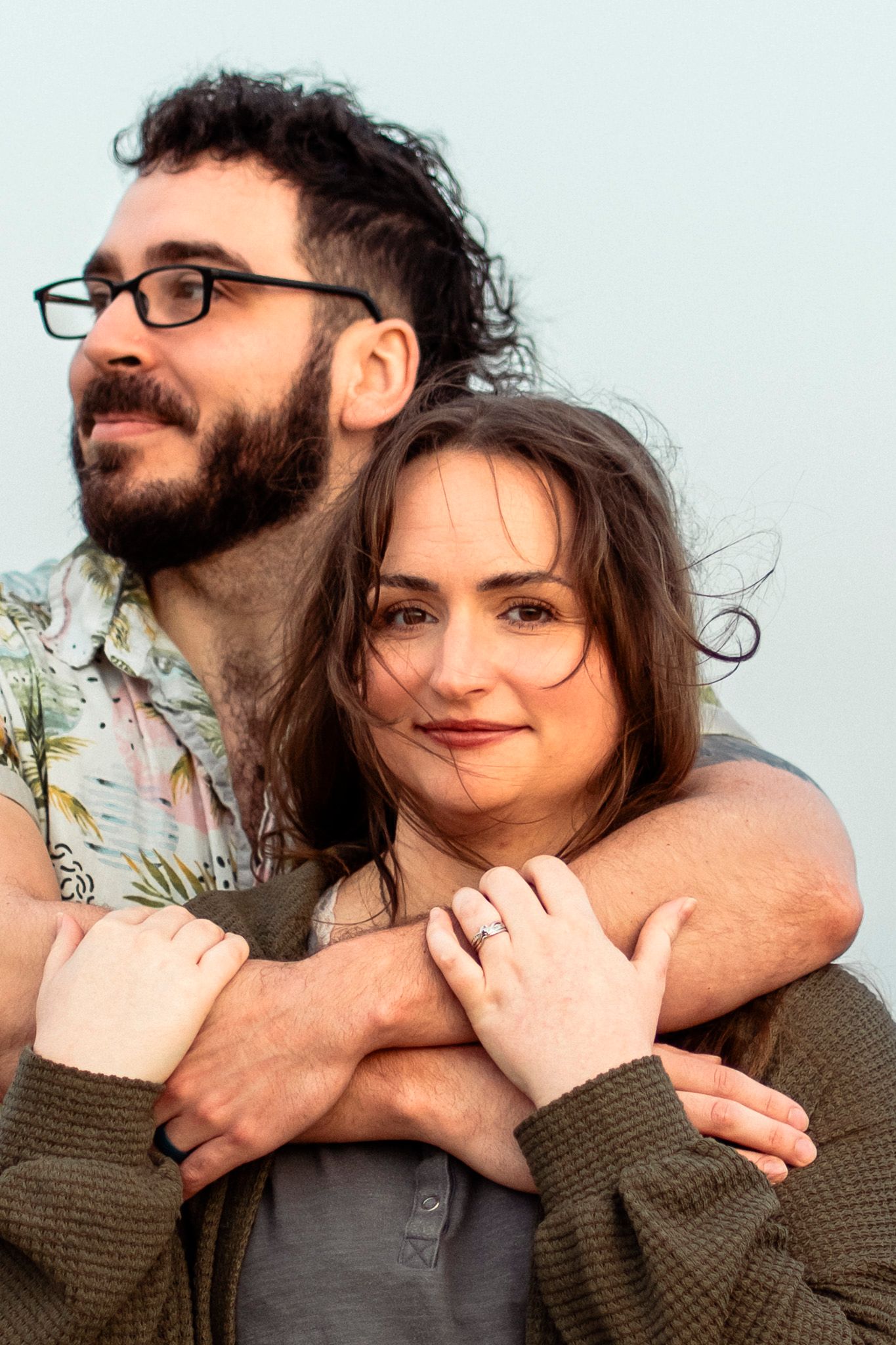 Couple embracing outdoors, woman showing engagement ring while man wraps arms around her.