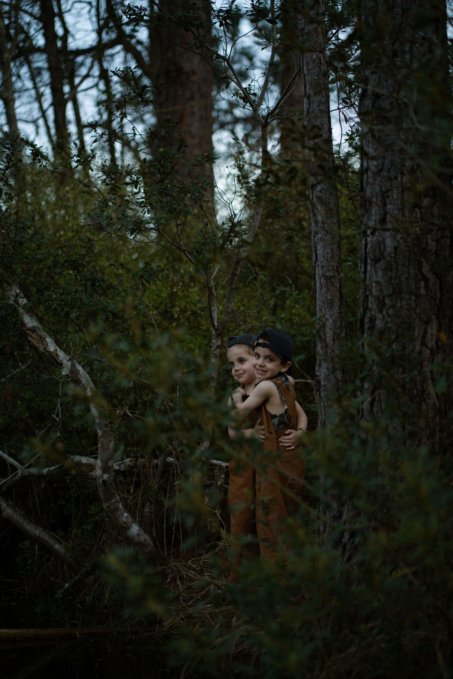 Two children embracing among dense green pine trees in a dark moody forest setting.