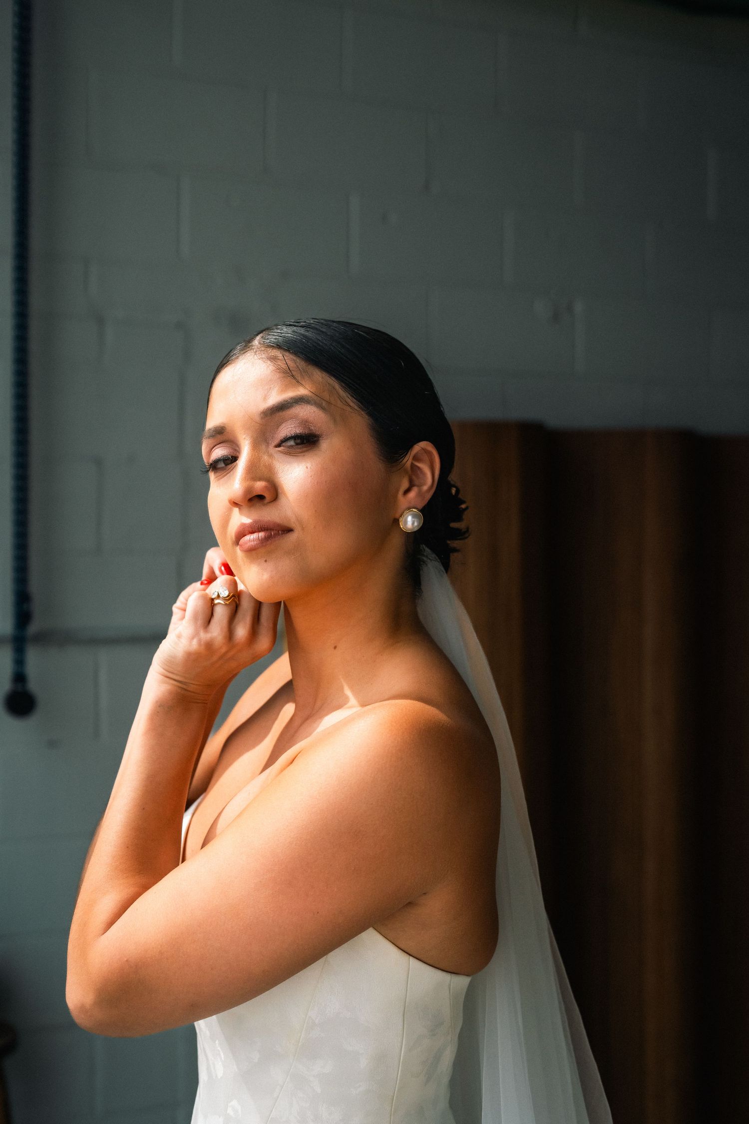 A bride in a strapless white gown and veil touches her pearl earring, posing thoughtfully against a dark wall.