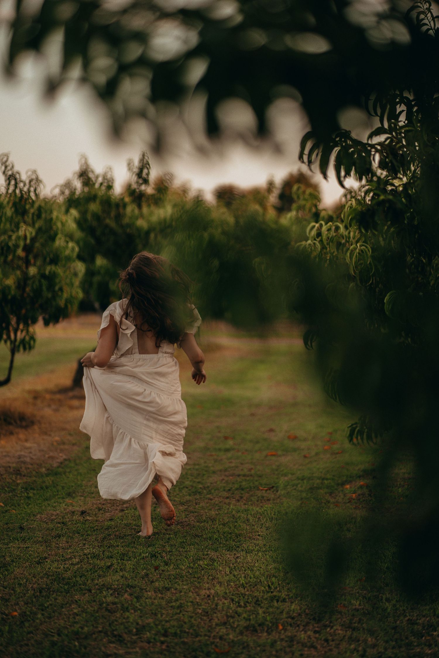 Young girl in white dress running barefoot through green orchard pathway at dusk.