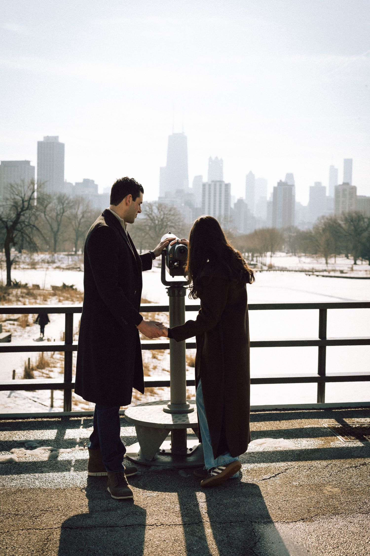 Two silhouetted figures stand at a snowy observation point overlooking a city skyline on a winter day.