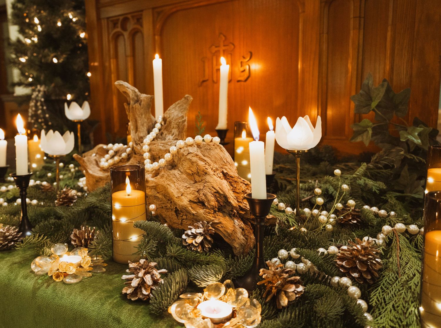 Elegant Christmas tablescape with white candles, pine cones, greenery, driftwood, and pearl garlands in warm candlelight.