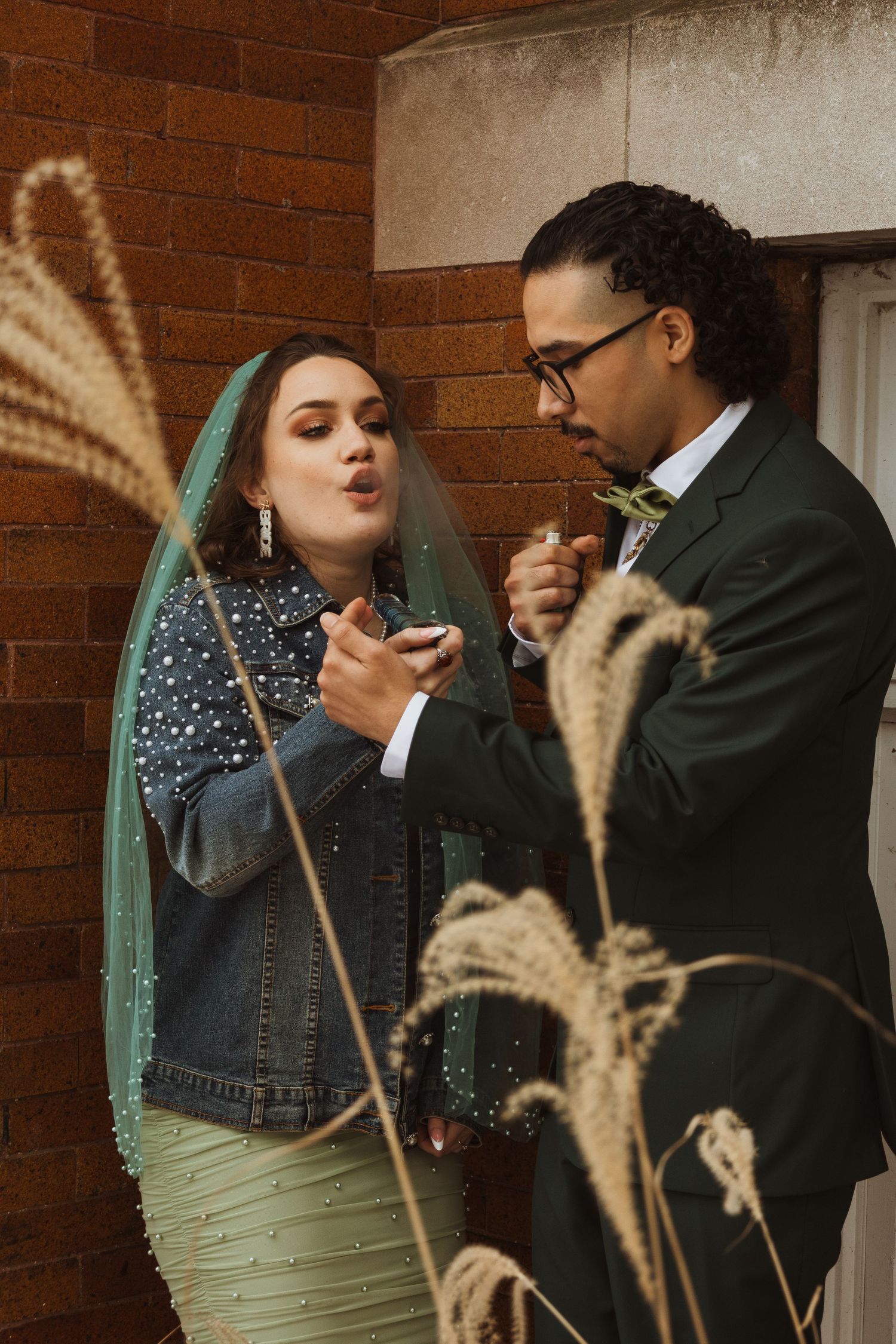 Bride in green veil and groom in dark suit lighting cigarettes outside brick building.