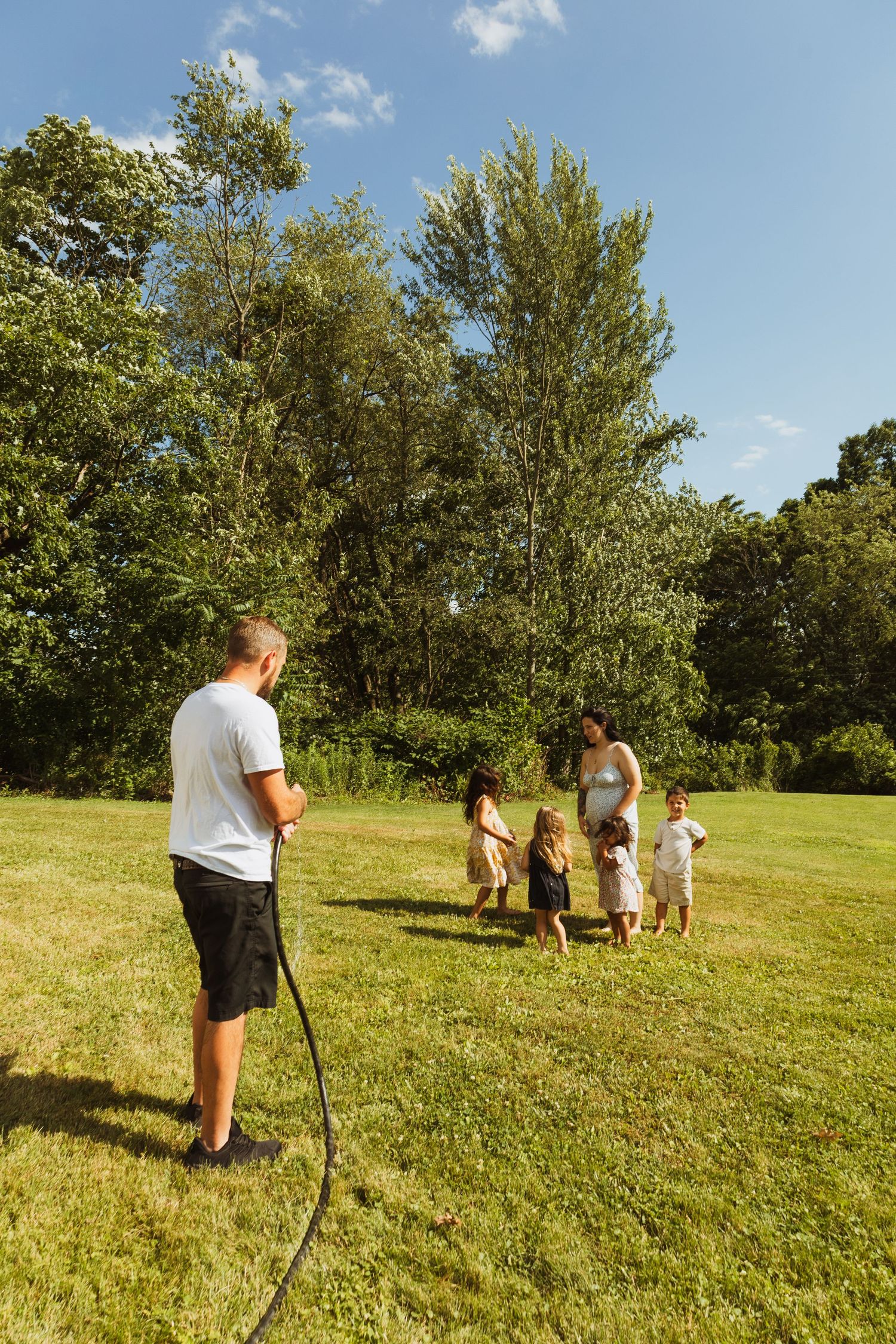 Family playing with garden hose on sunny green lawn surrounded by trees on a beautiful summer day.