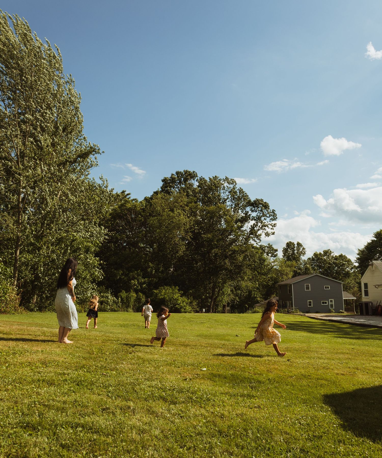 Children playing and running on a lush green lawn surrounded by trees and a house on a sunny day.