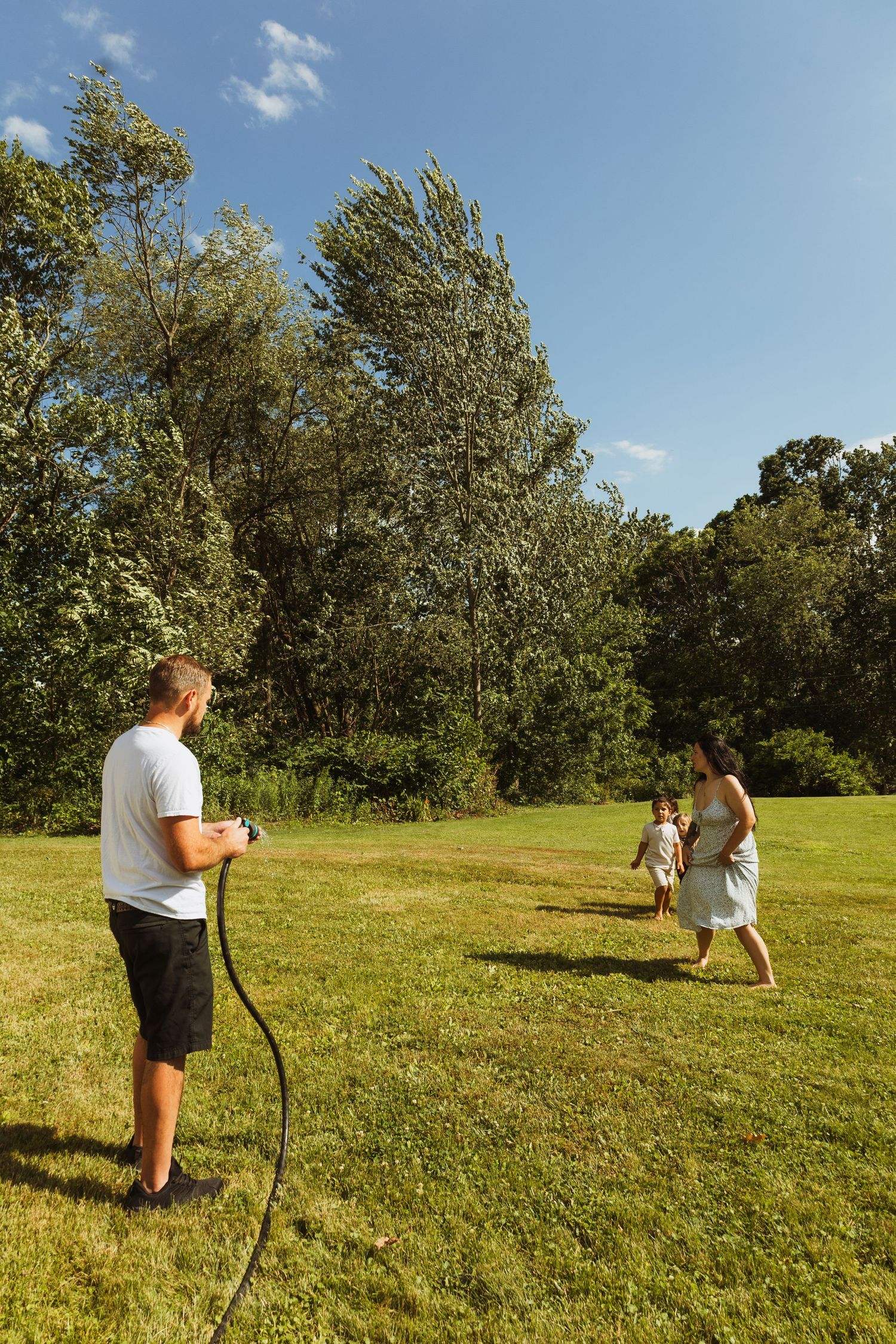 Family enjoying a sunny day outdoors, man holding garden hose while woman and child play on green lawn.