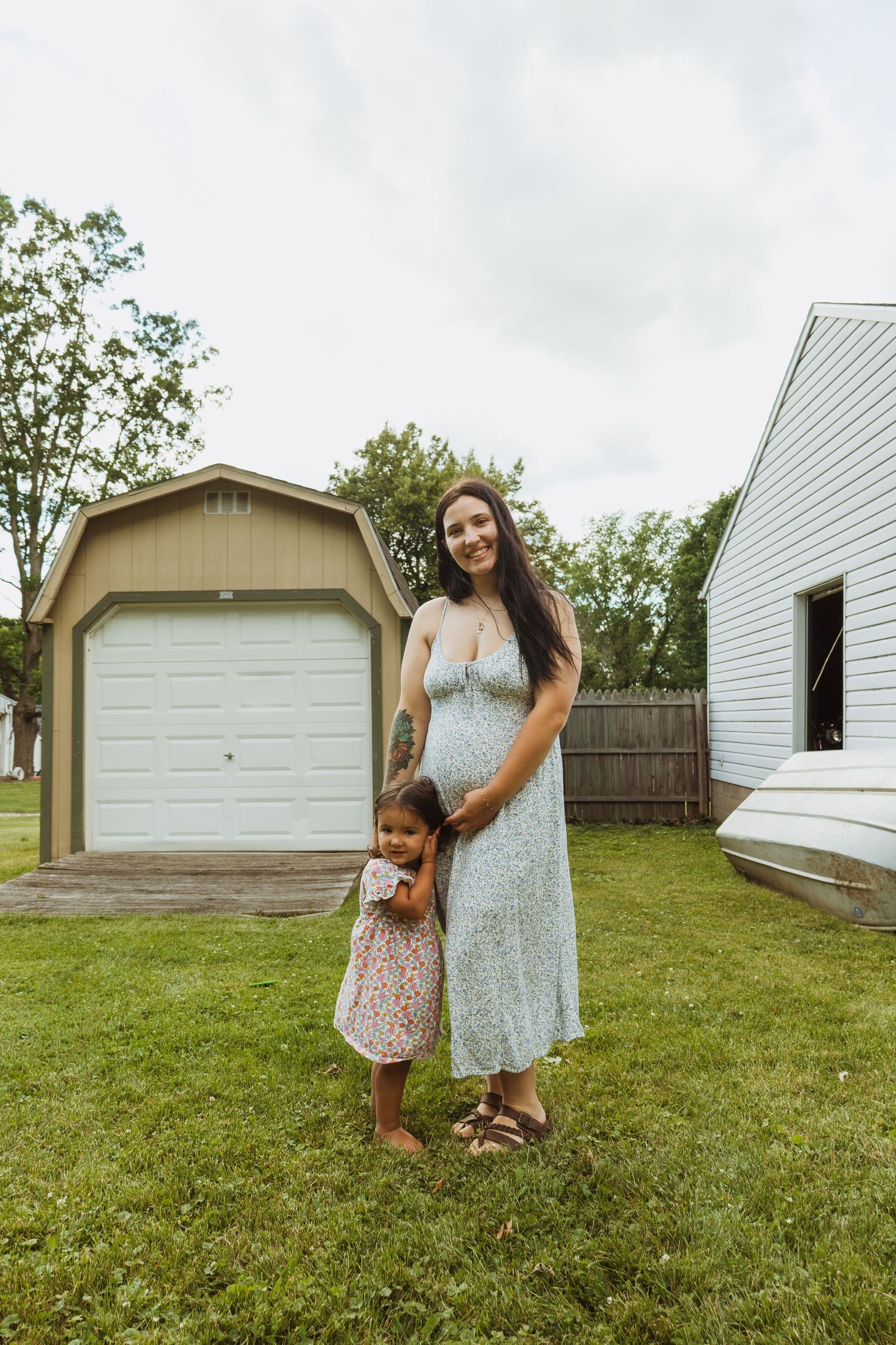 Pregnant woman in floral dress smiling with young daughter in backyard near garage on cloudy day.