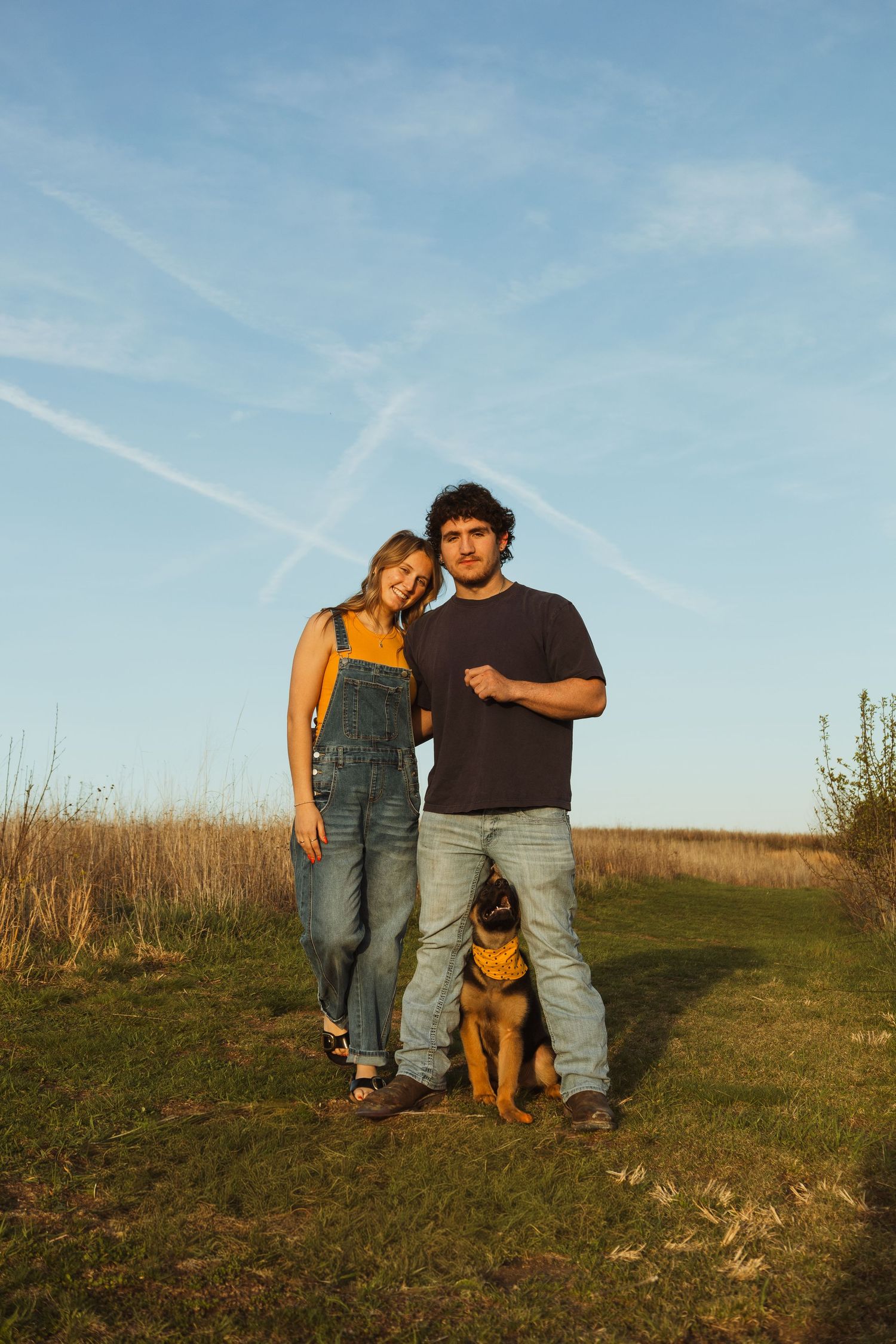 Young couple standing outdoors in a field with their Rottweiler dog under a blue sky at golden hour.