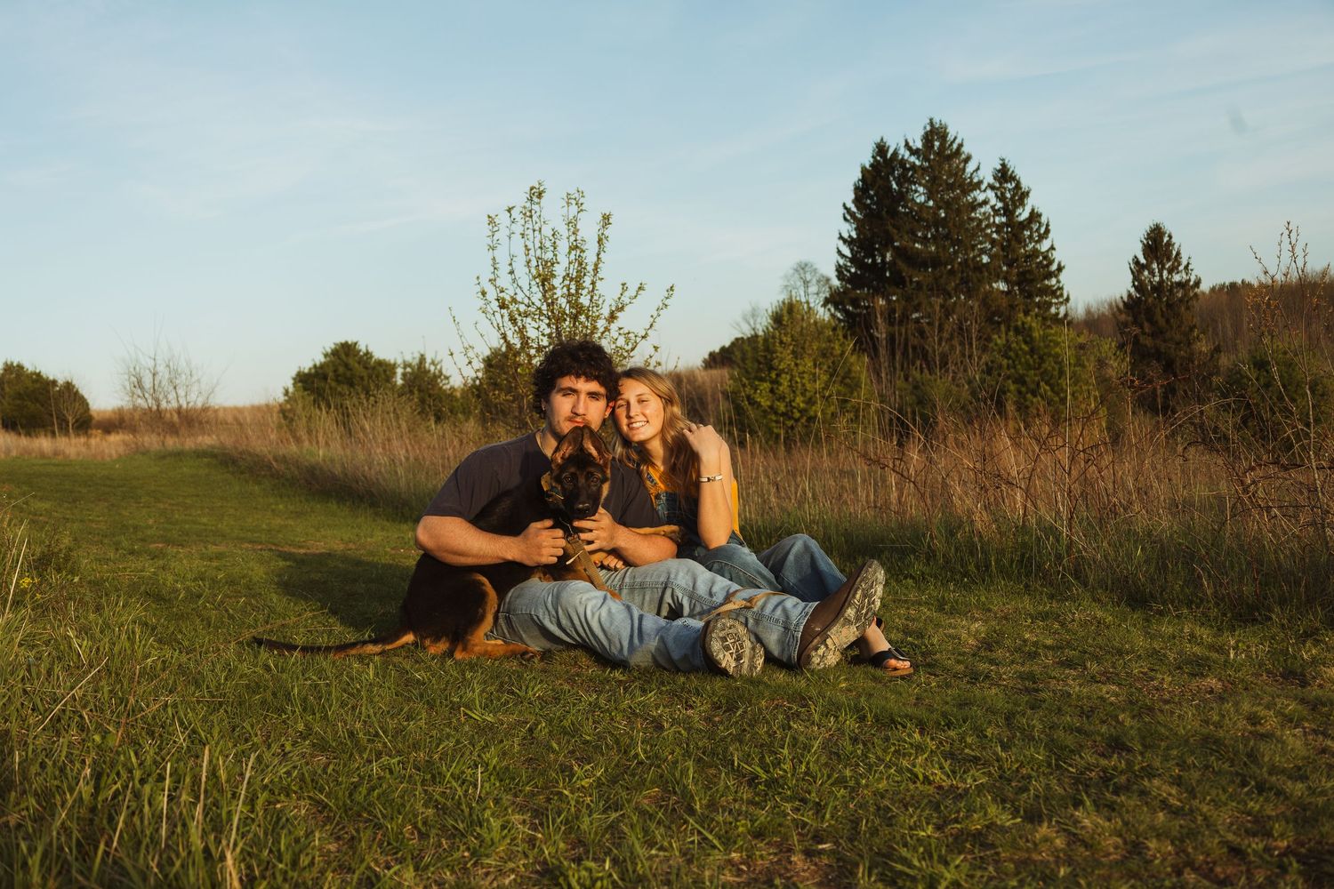 Couple sitting together on green grass field with their dog, surrounded by trees and tall grass in warm golden sunlight.