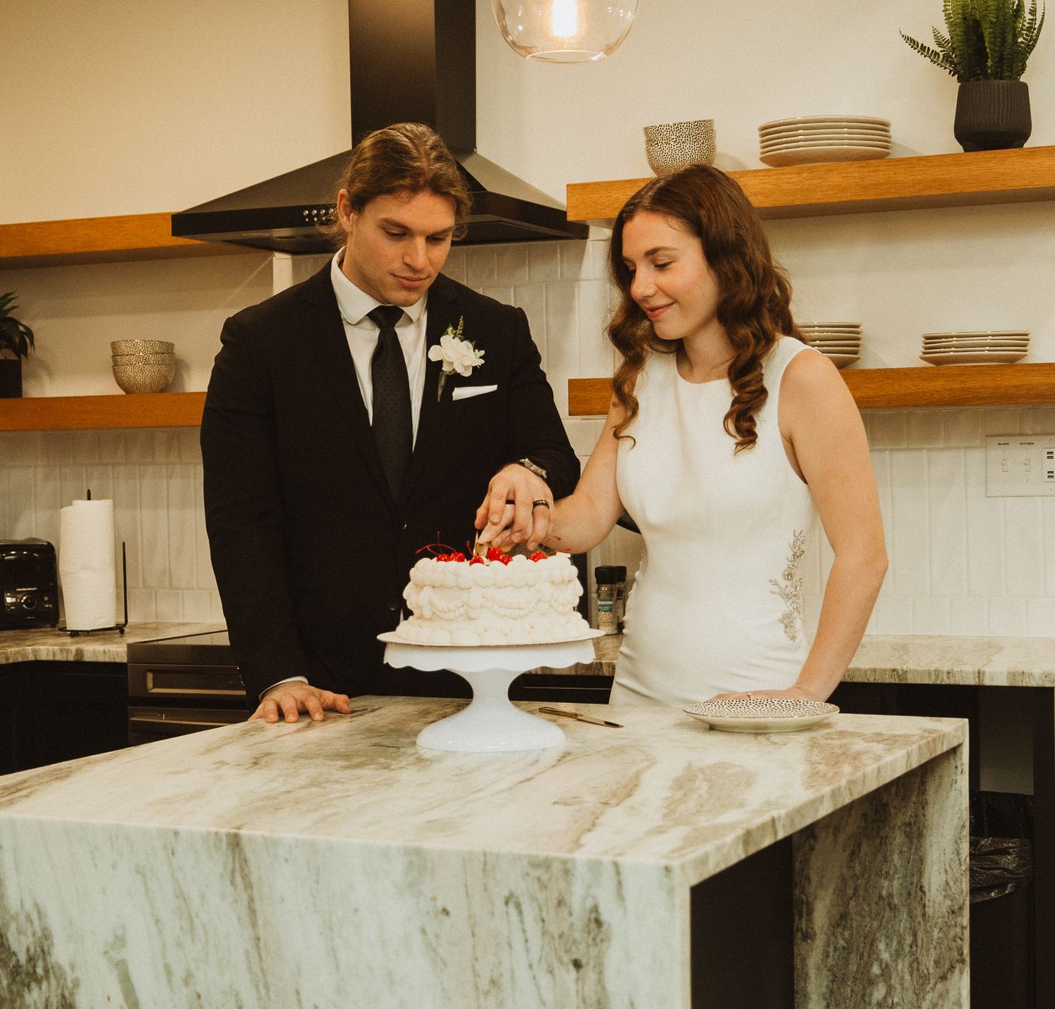 A couple cuts their elegant white wedding cake together on a marble counter.