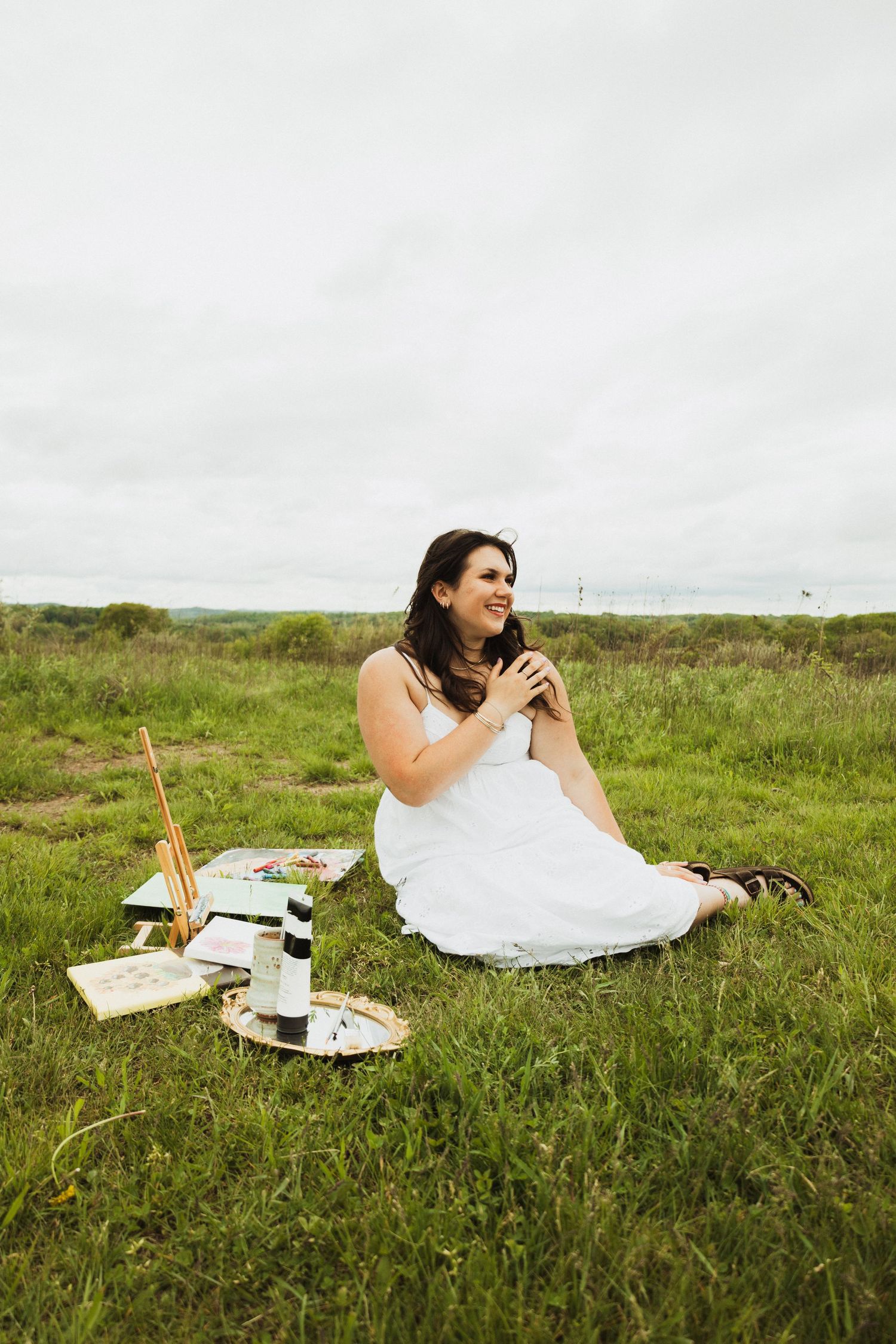 A person in a white dress enjoys a romantic picnic on a grassy hill with wine and snacks laid out on a blanket.