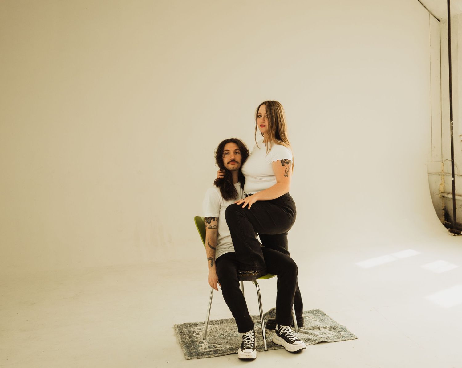 Artistic studio portrait with two people posed on chair against white backdrop.