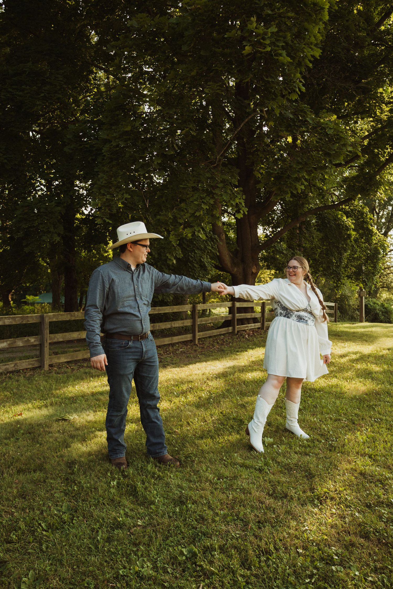 Playful dance scene on grassy hillside with wooden fence and trees in golden sunlight.