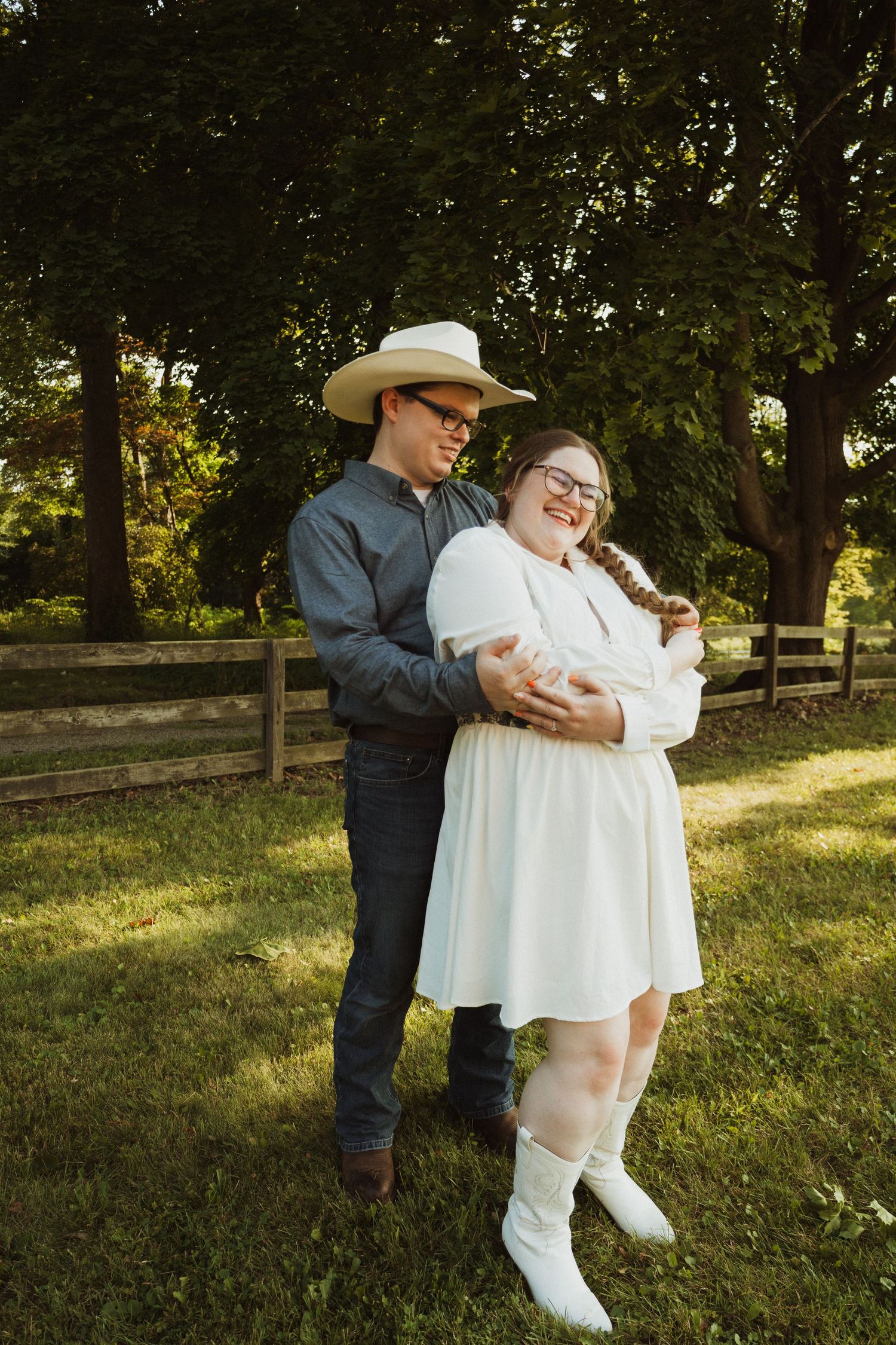 A couple embraces in a romantic pose outdoors with white boots and western attire against a leafy green backdrop.