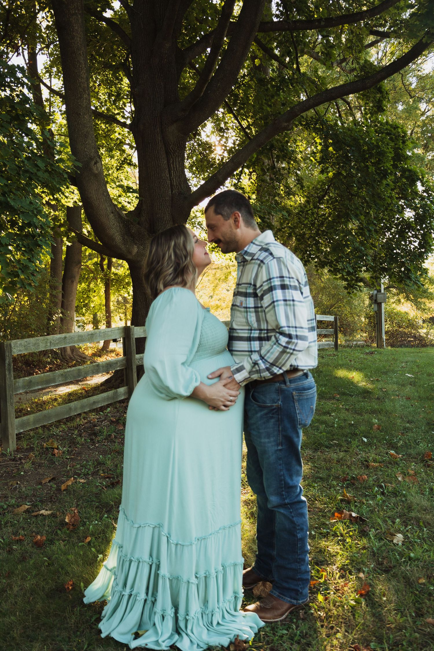 A couple in a romantic outdoor setting with trees and a wooden fence share tender moments in matching mint and plaid.