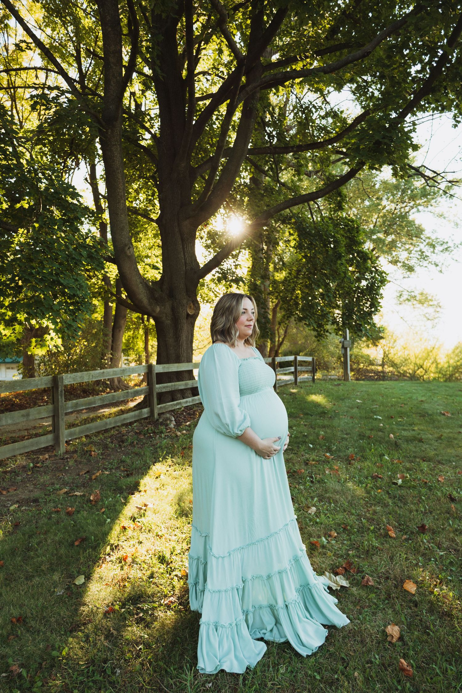 A couple in matching mint green dresses embrace on a grassy hillside during golden hour as sunlight filters through trees.