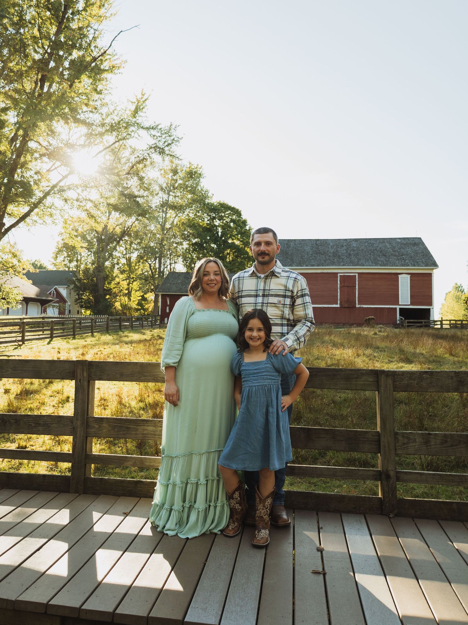 Family poses together on wooden deck at sunset with red barn and trees in background.