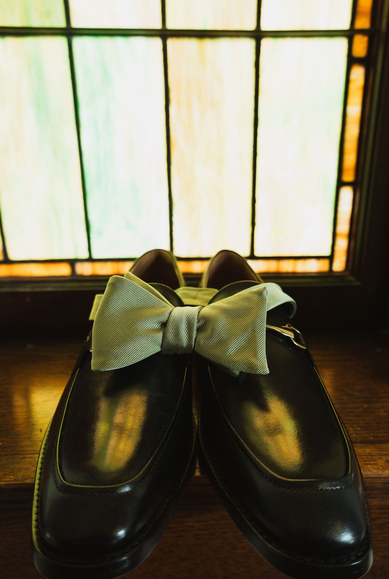 Black dress shoes with a sage green bow tie resting on top, displayed before a stained glass window.