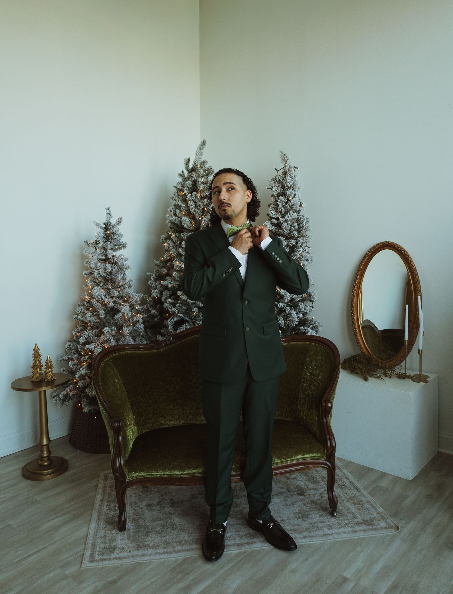 Groom in dark green suit adjusting bow tie beside flocked Christmas trees and vintage mirror.
