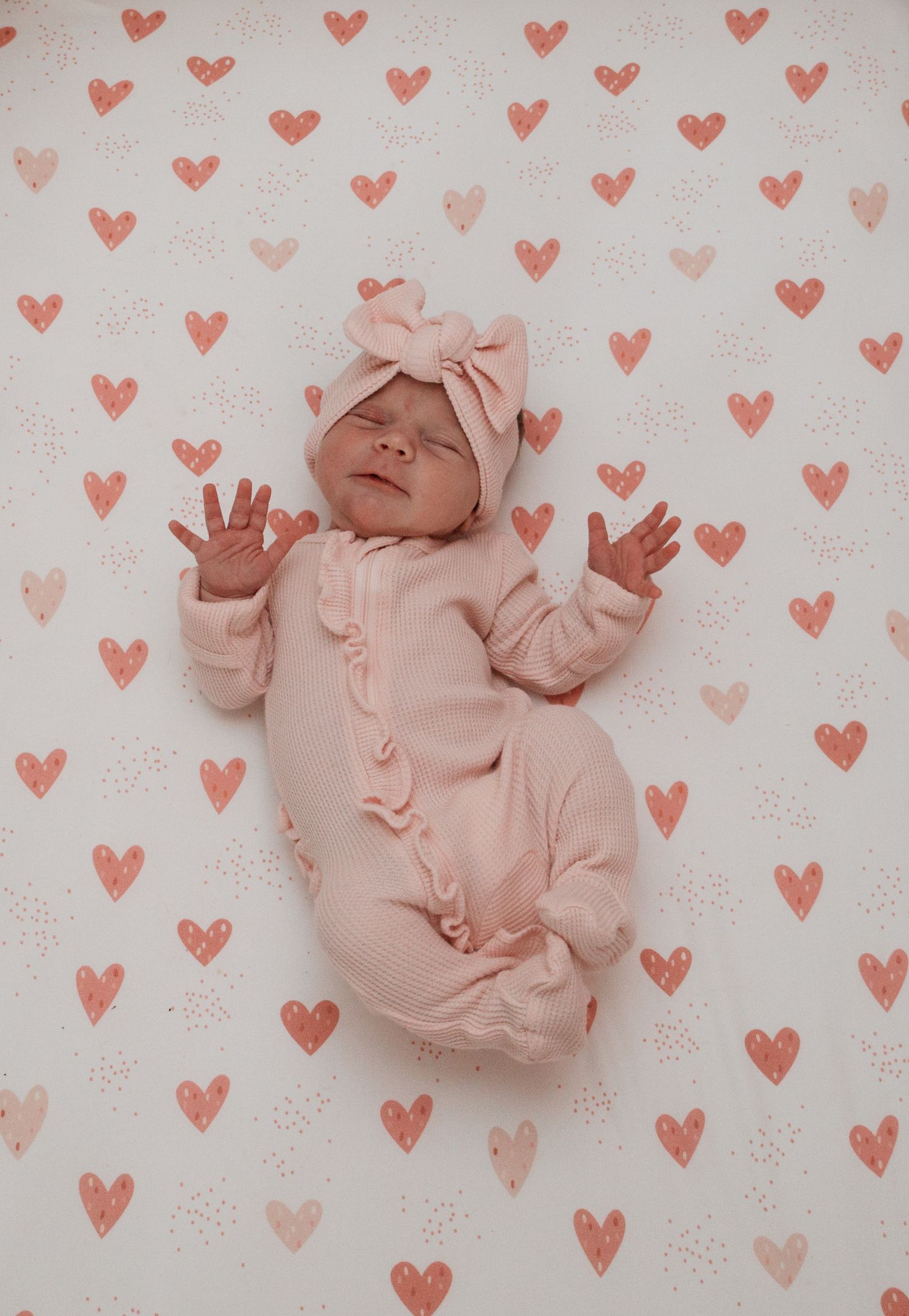 Sleeping baby in pink outfit and matching bow lying on heart-patterned background in soft pink tones.