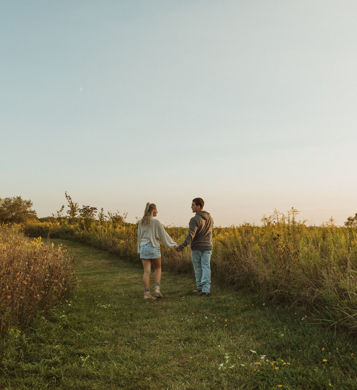 Couple holding hands while walking through scenic meadow path at dusk.