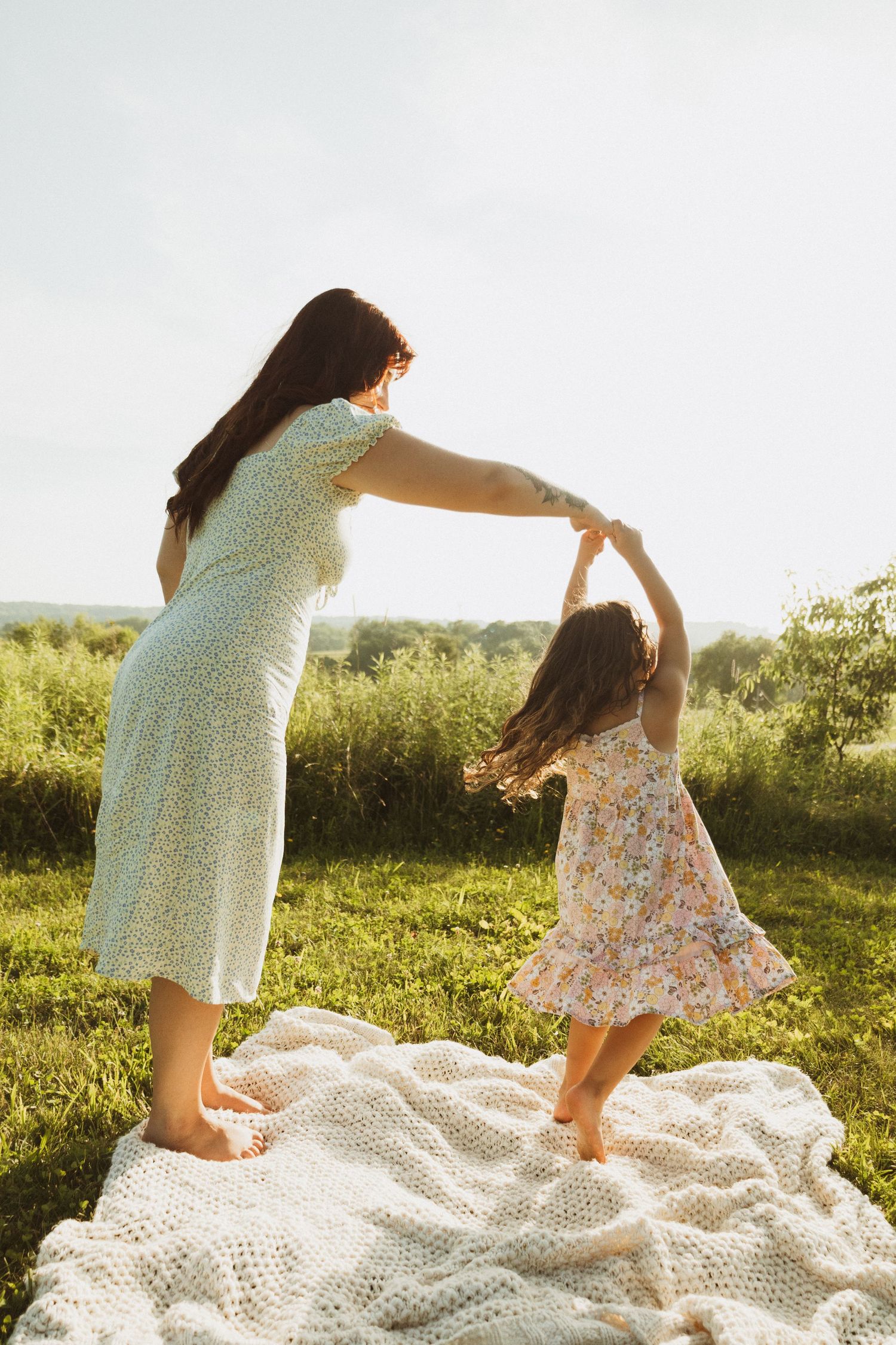 Two people in floral dresses hold hands while dancing together on a blanket outdoors.