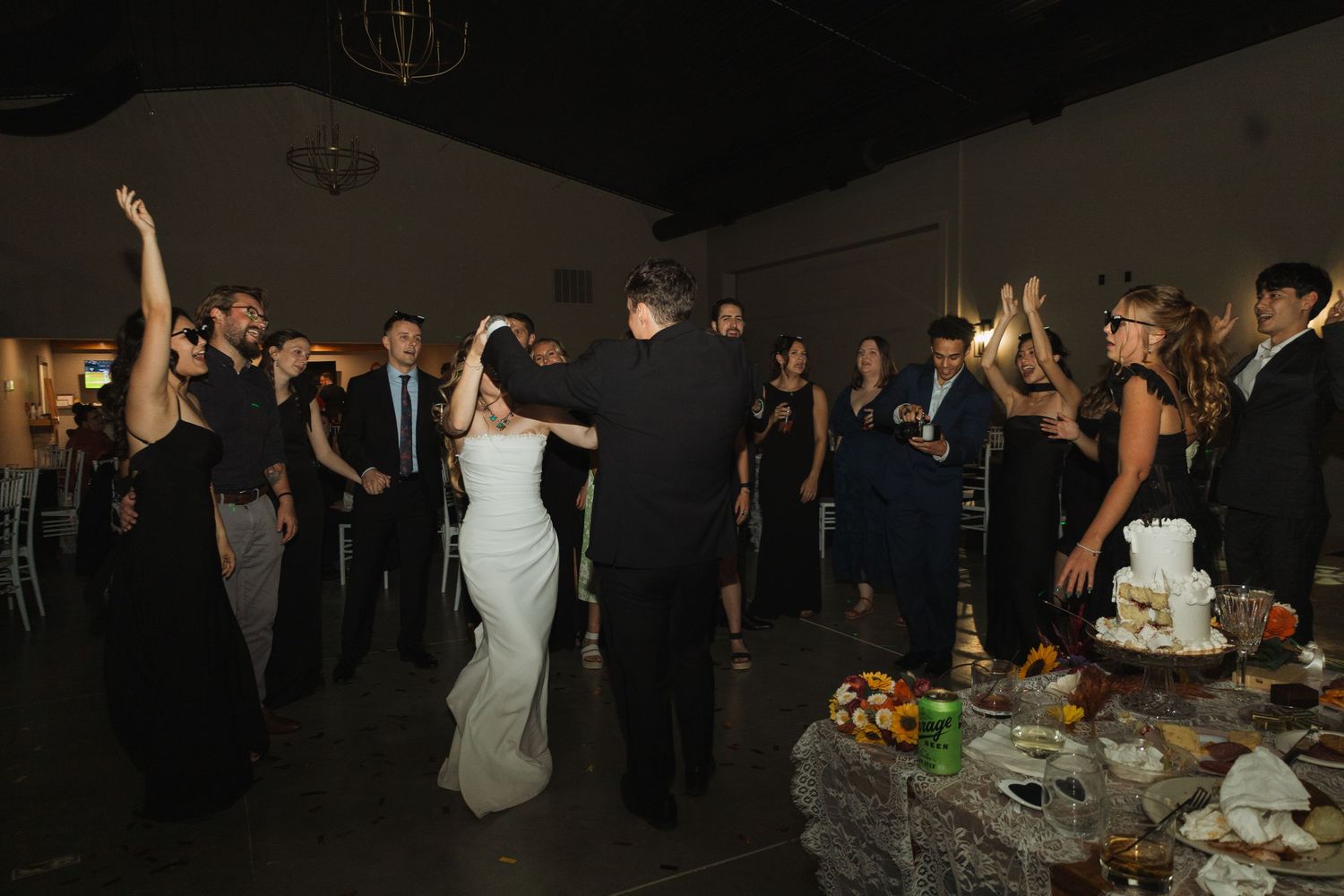Wedding guests dance and celebrate at a reception with a wedding cake displayed on a decorated table in a dark venue.