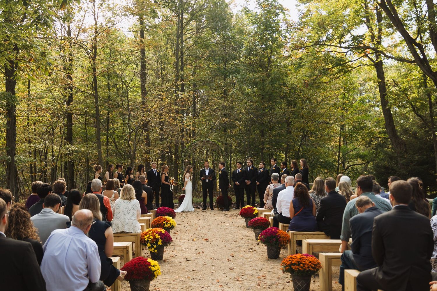 Outdoor wedding ceremony setup in a wooded area with autumn foliage and red floral arrangements.