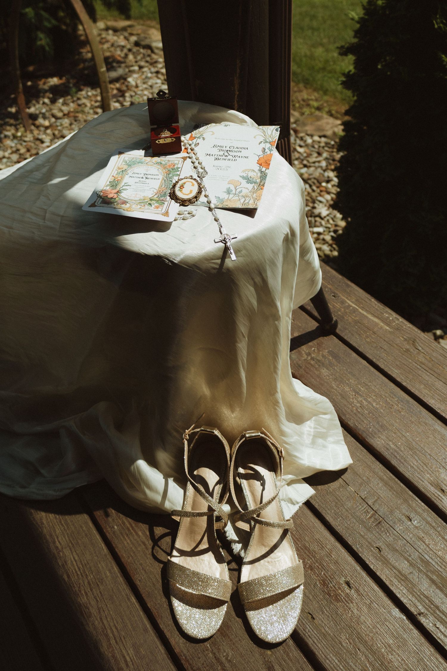 Wedding accessories including invitations and sparkly sandals arranged on wooden deck in sunlight.