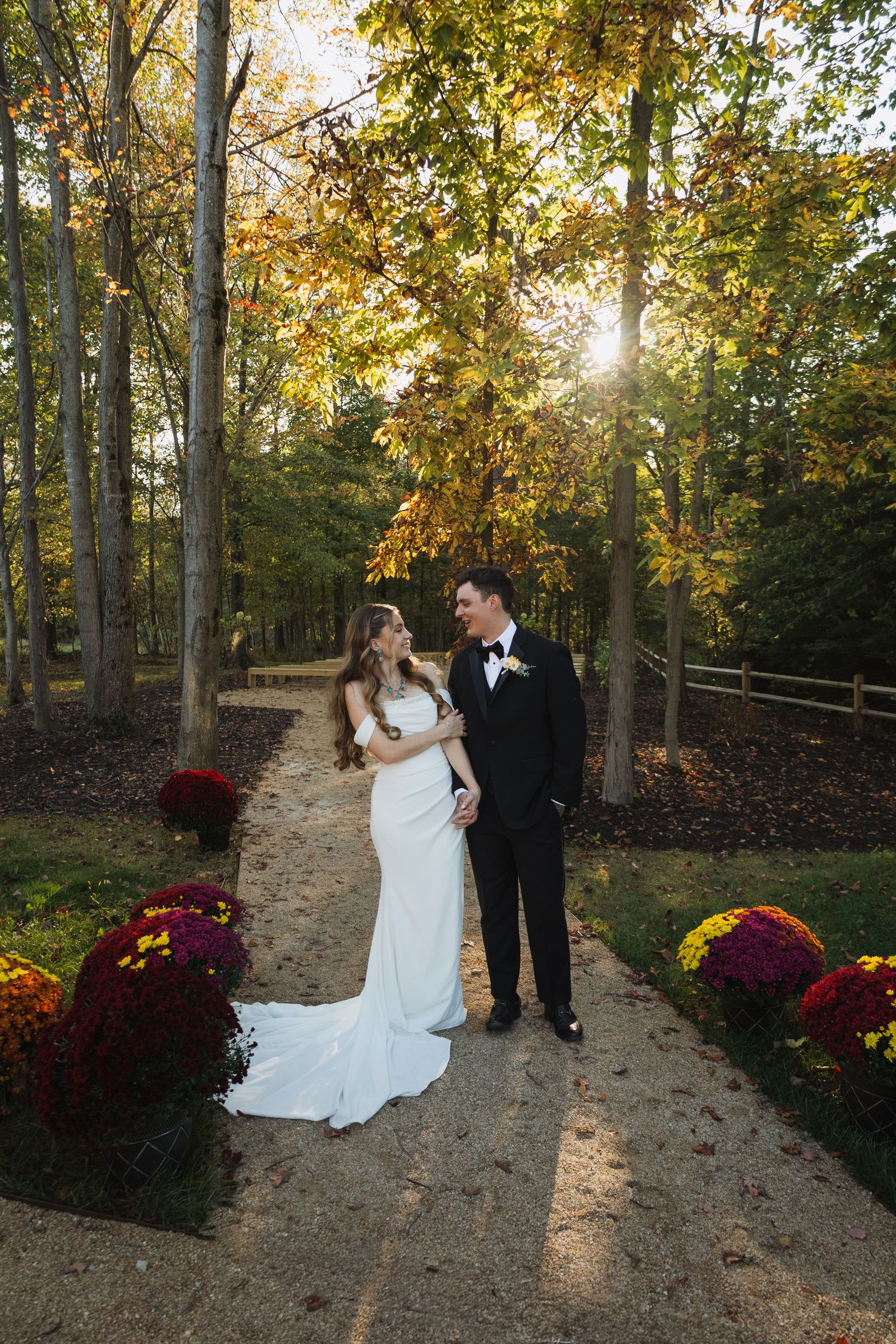 Couple poses on outdoor path surrounded by autumn trees and colorful mums in full bloom.