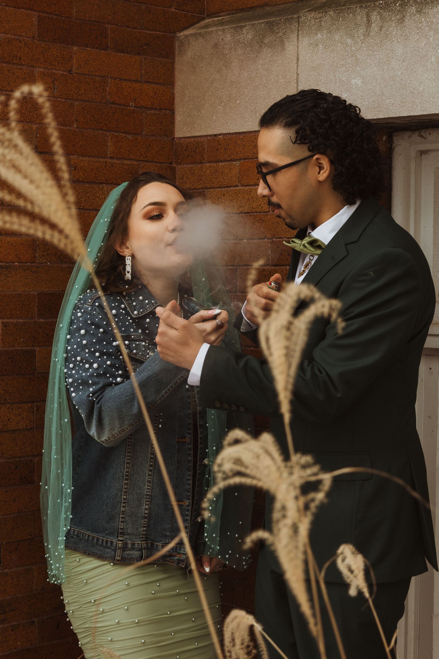 Bride exhaling smoke while groom lights cigarette outside brick building, both in wedding attire with teal veil.