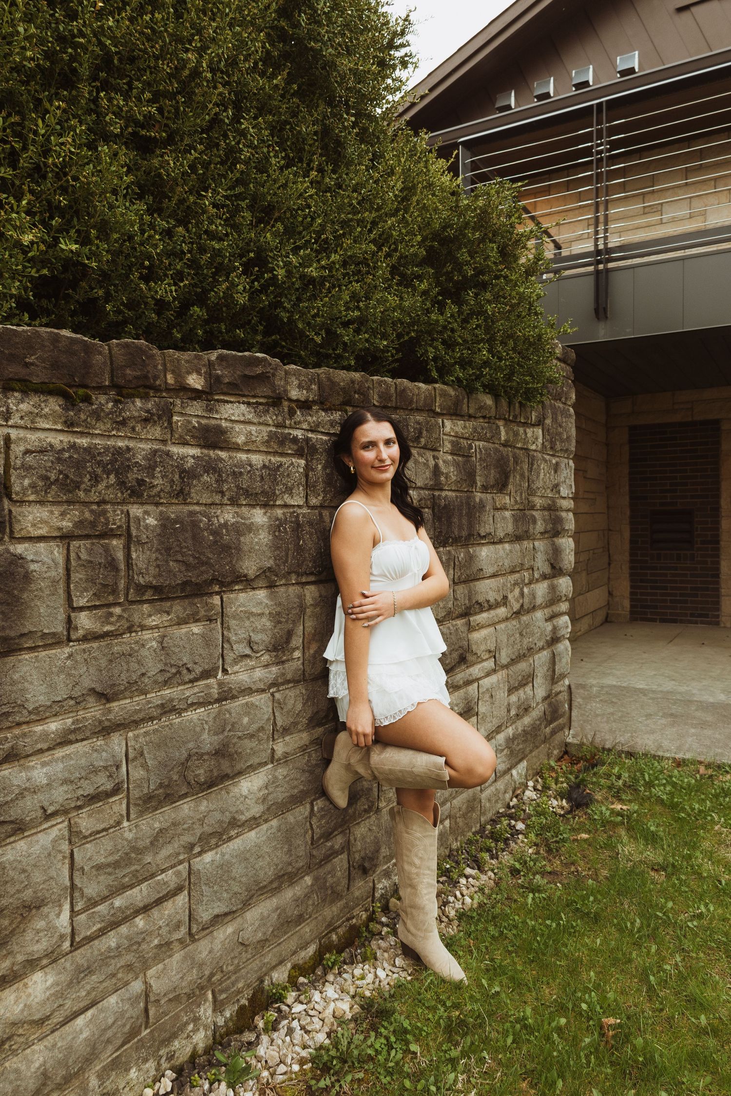 Young woman in white dress and cowboy boots leaning against a stone wall outdoors.