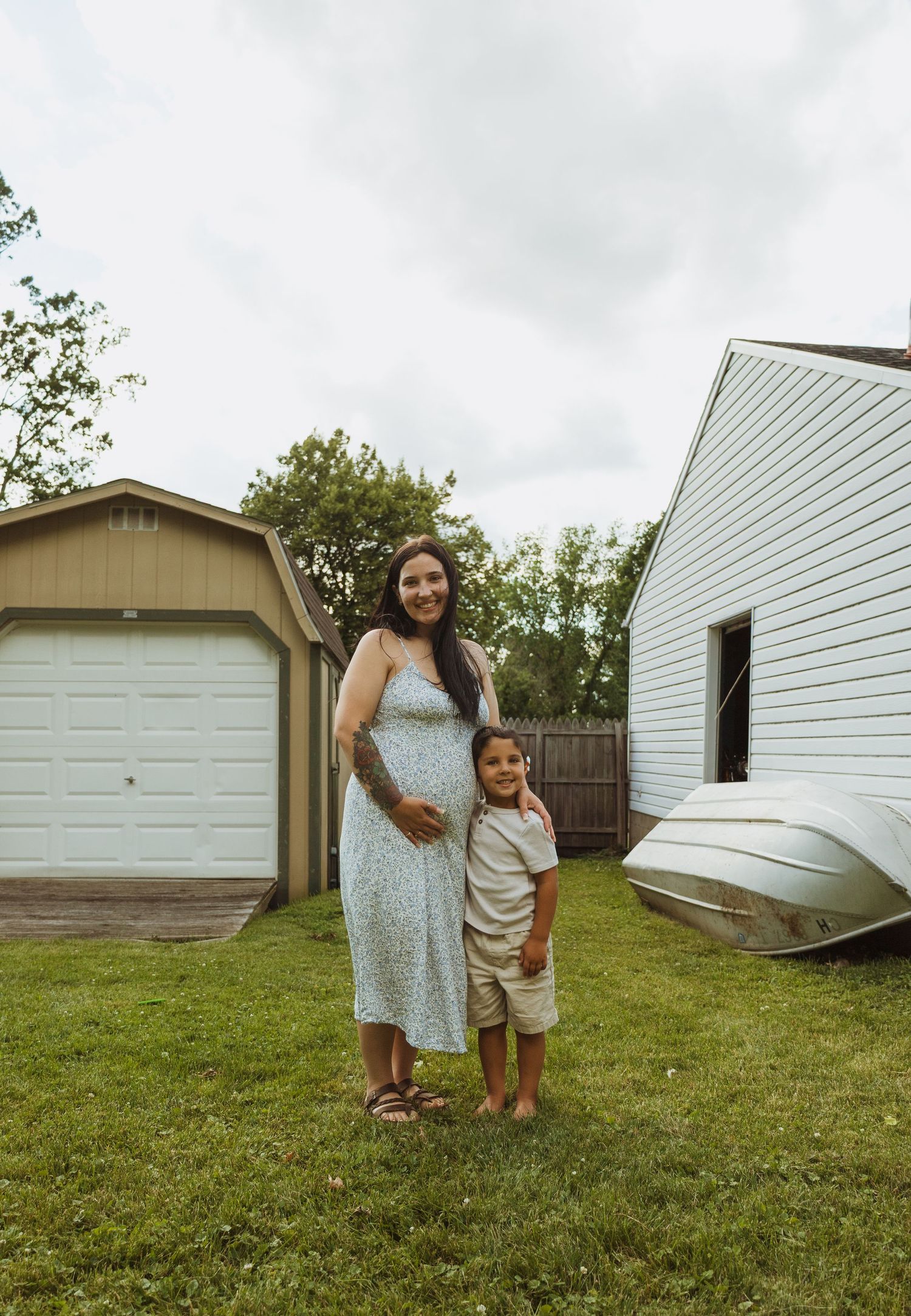 Pregnant woman in floral dress poses with young boy on green lawn beside garage and white building.
