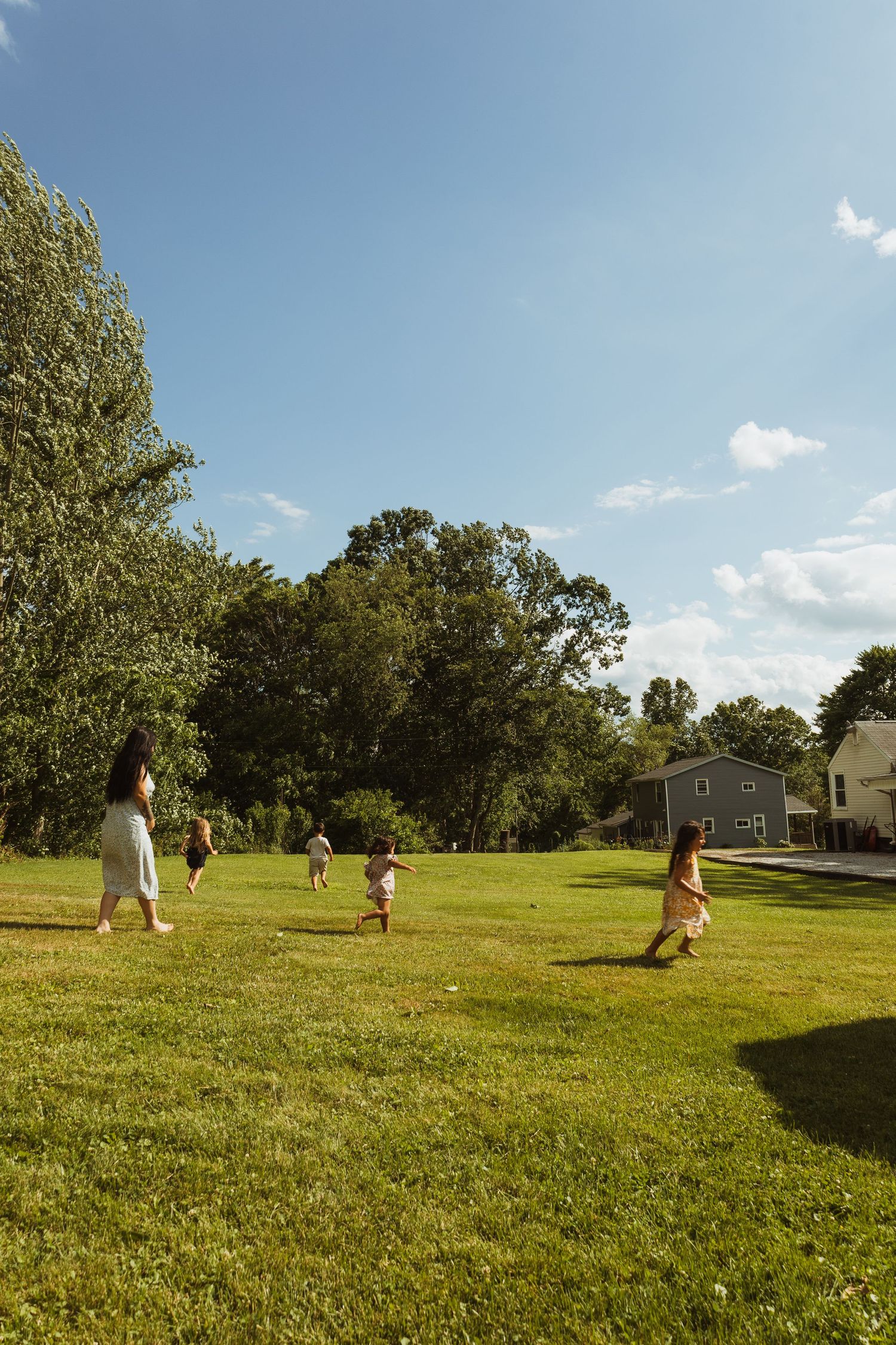 A family plays together on a lush green lawn surrounded by trees and a house under a bright blue sky.