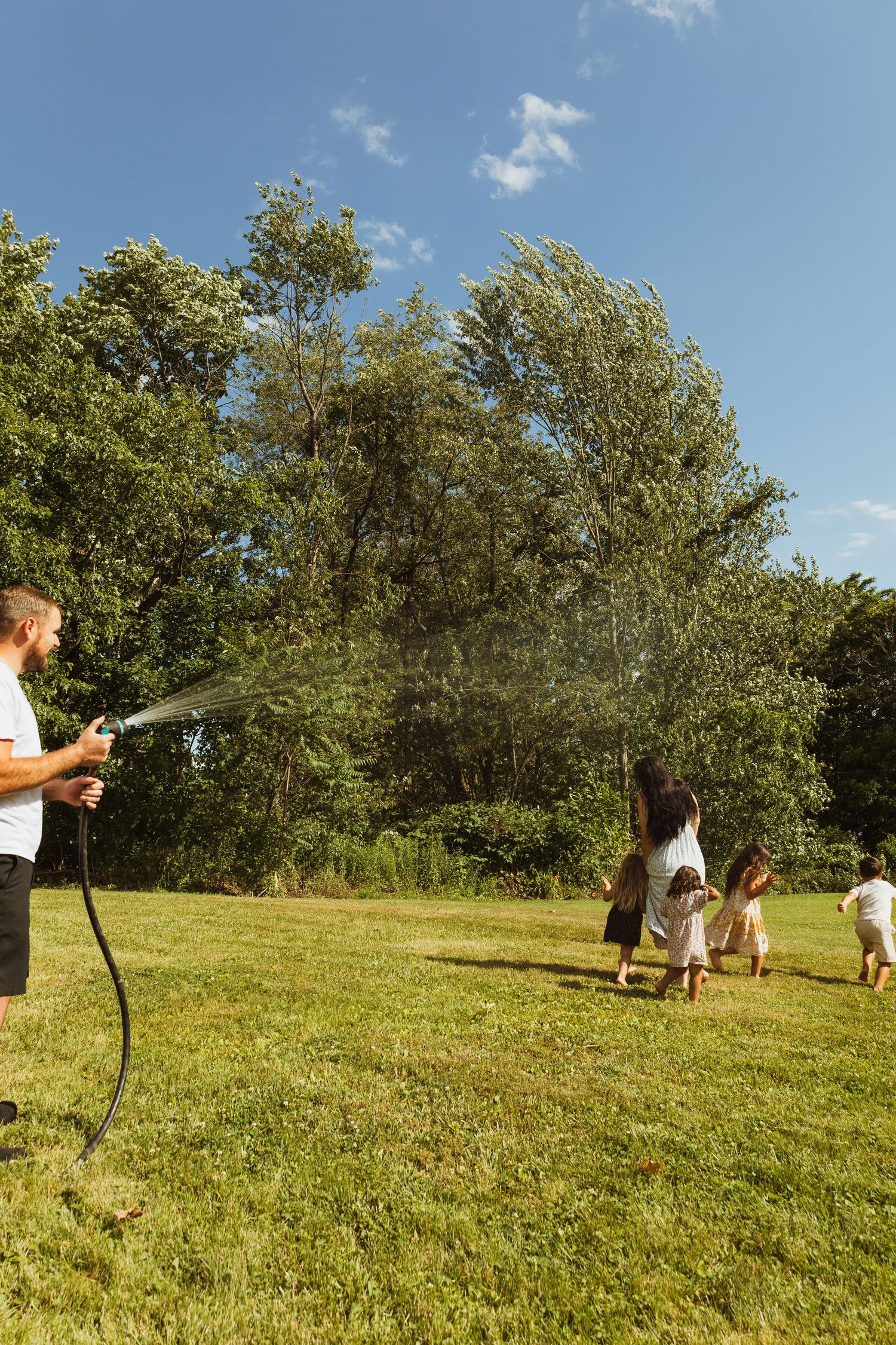 Family playing with garden hose on sunny day, father spraying water while children and mother run across green lawn.
