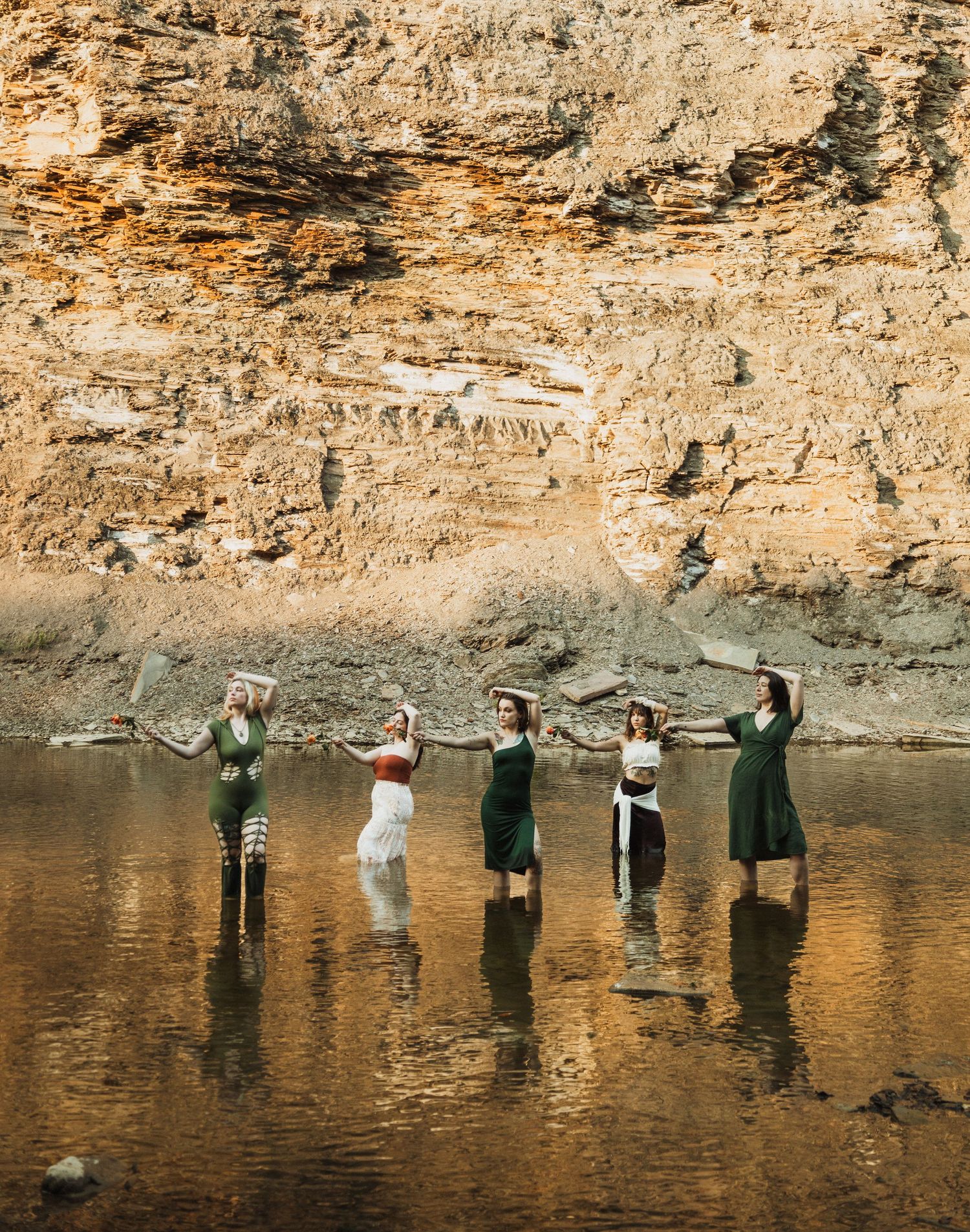 Group of people standing in a line in shallow water with their arms raised against a tall cliff wall.