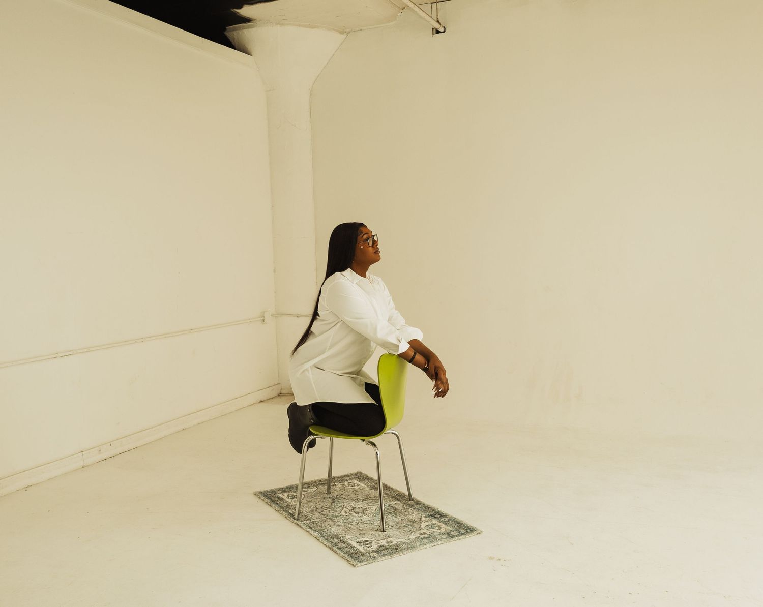 A person in white clothing sits sideways on a chair placed on a decorative rug in a minimalist cream-colored room.