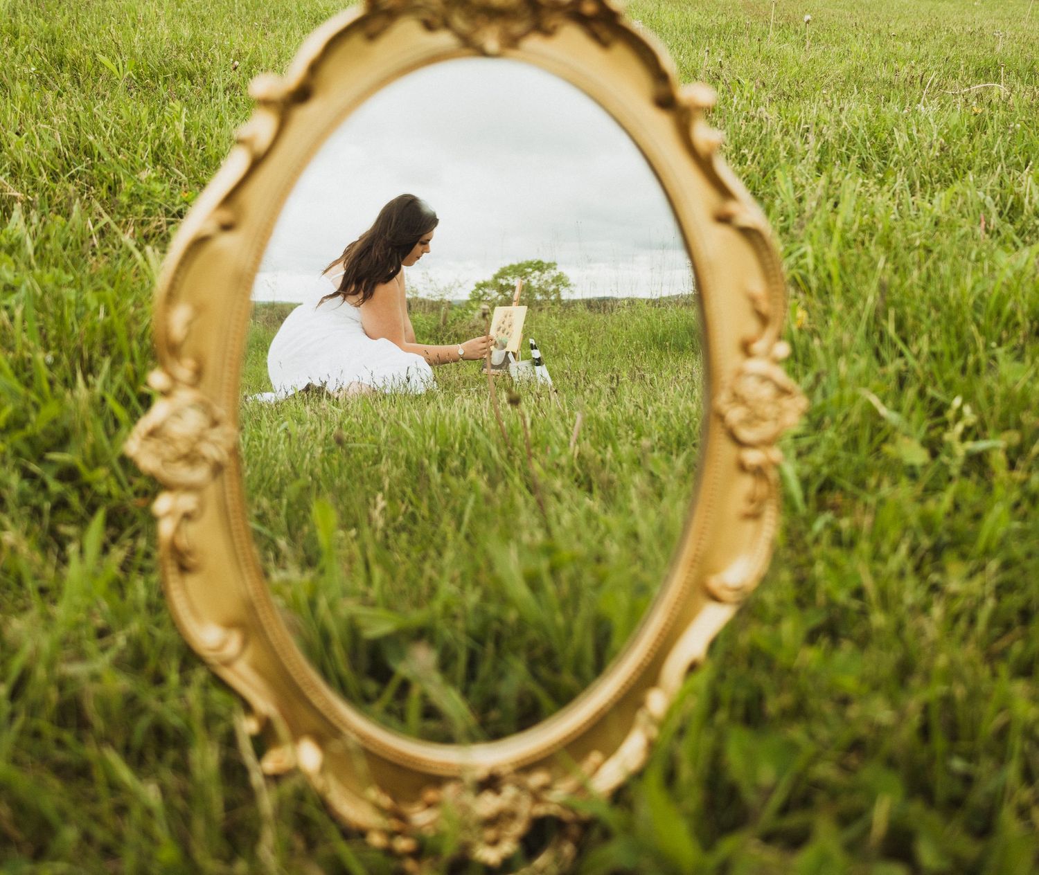 An ornate gold vintage mirror frame showcases a reflection of someone sitting peacefully in a green grassy meadow.