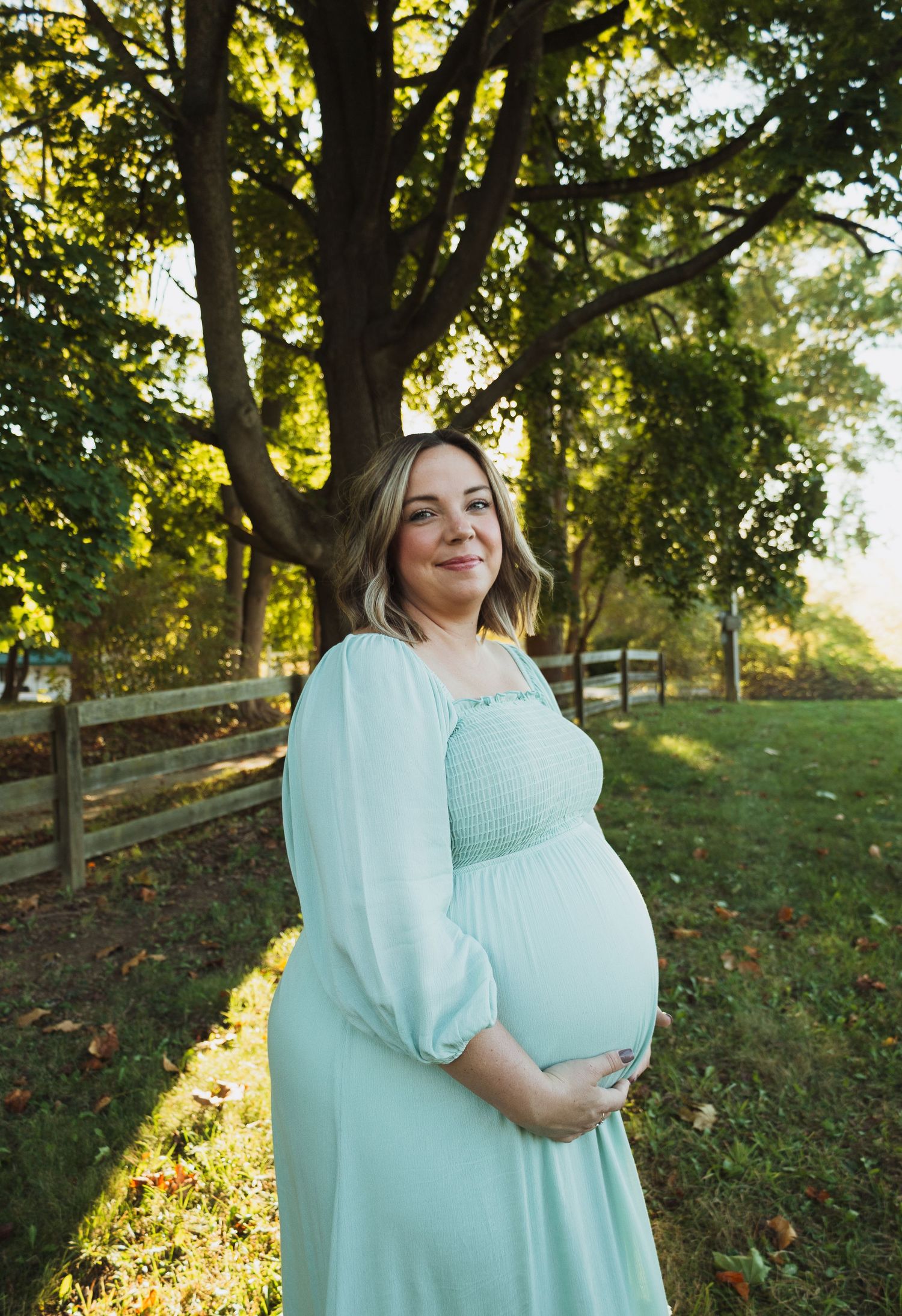 A person in a mint green maternity dress poses outdoors in golden sunlight with trees and a wooden fence in the background.
