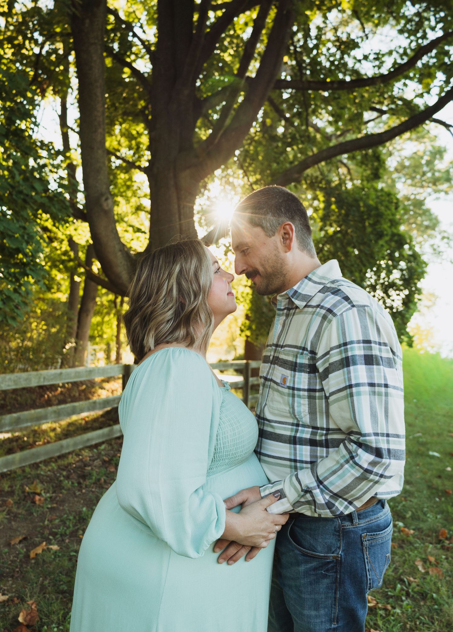 A romantic couple shares an intimate moment at sunset in a wooded park setting, with sunlight streaming through the trees.