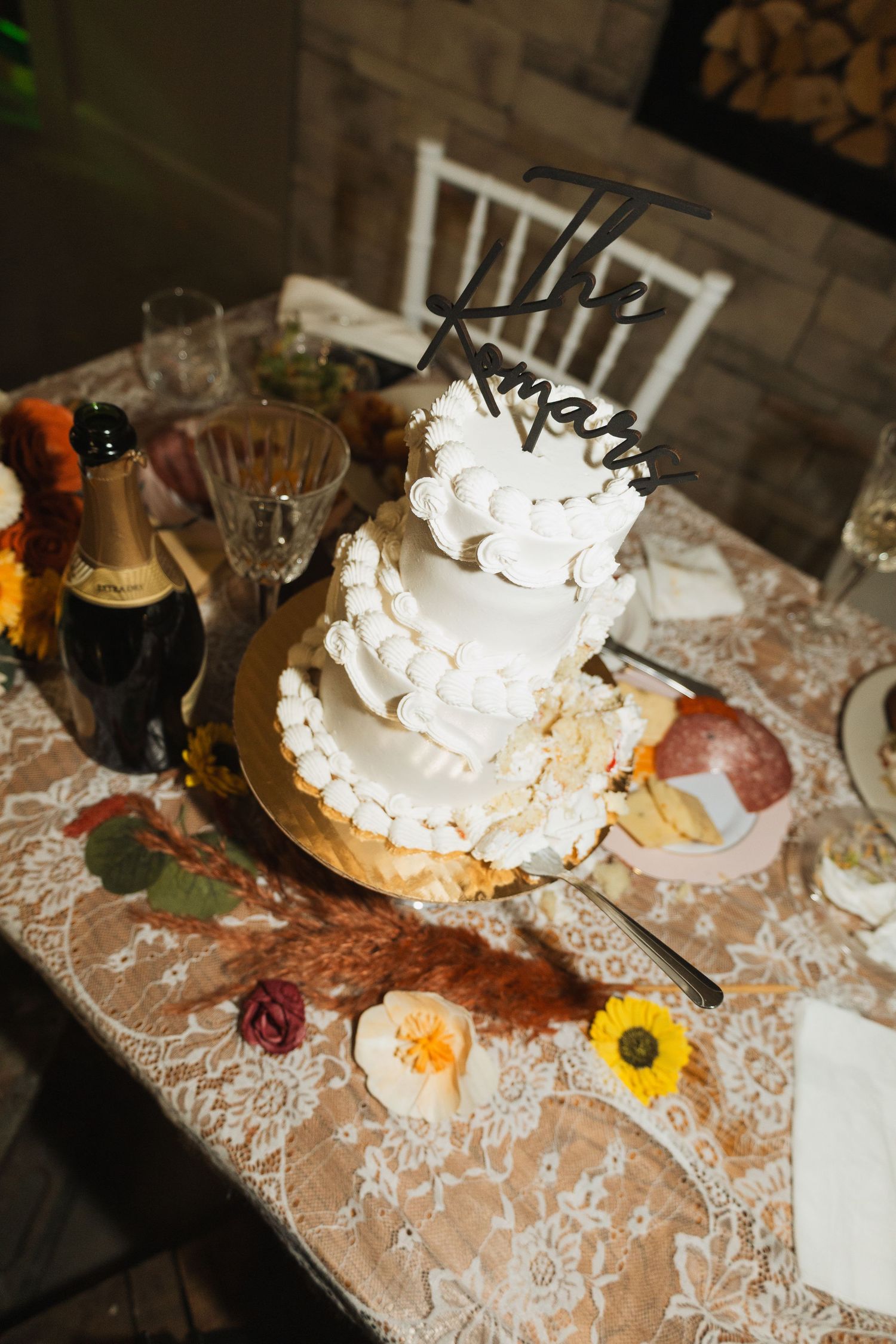 An elegant white wedding cake with ruffled tiers displayed on a lace tablecloth with floral decorations.
