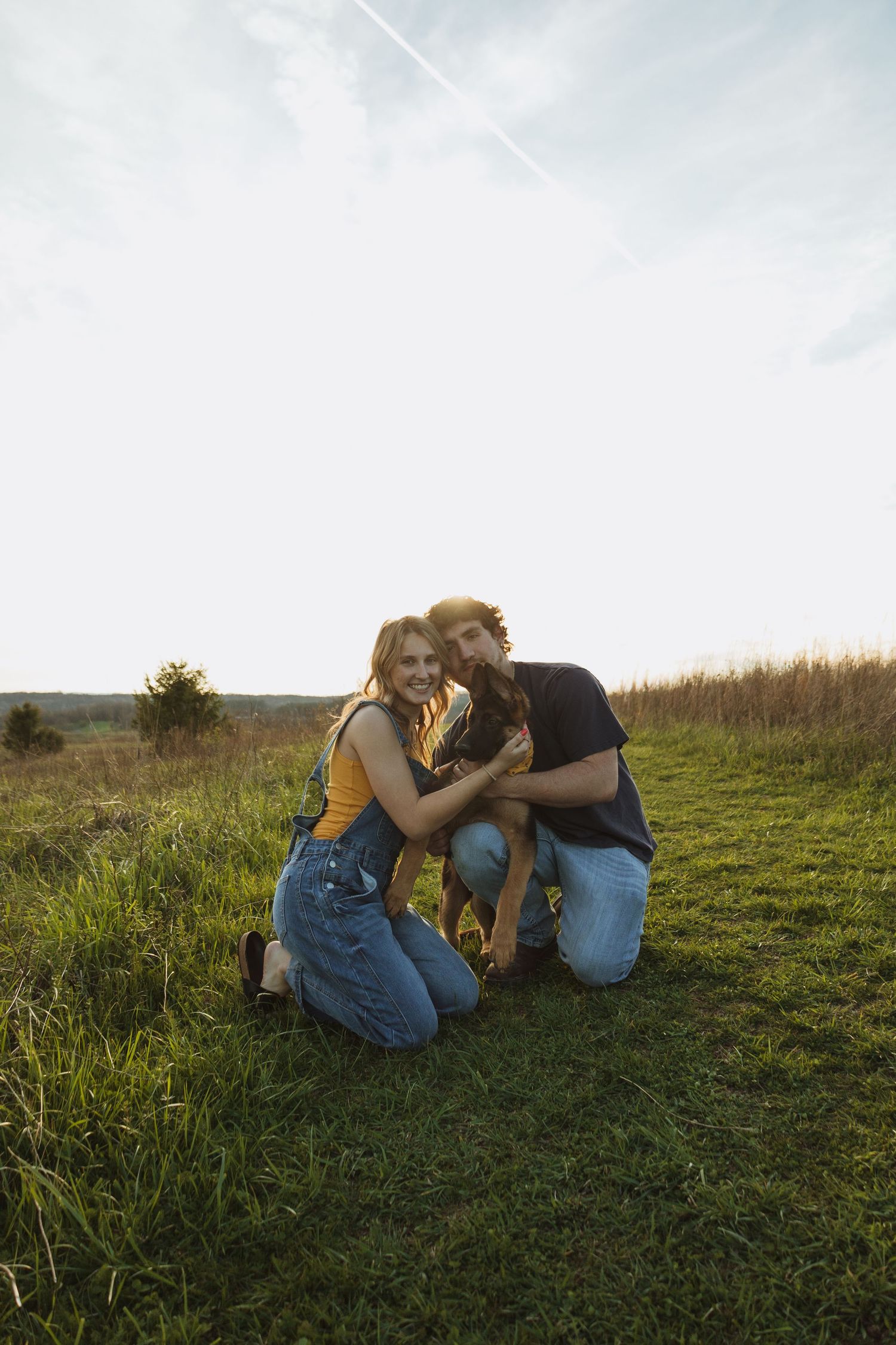 A smiling couple kneels together on green grass in a sunny field, petting their dog during golden hour.