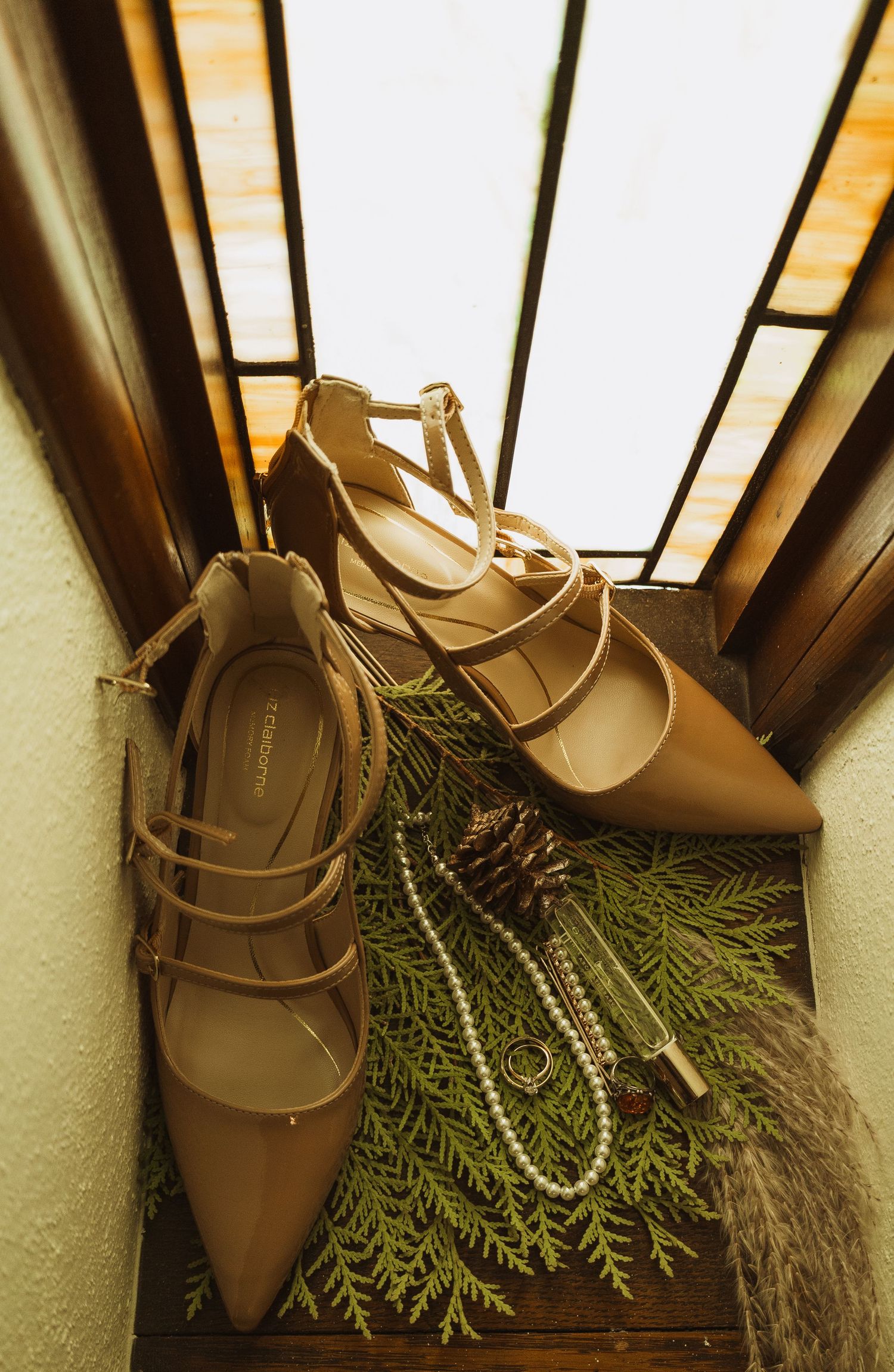Nude strappy heels with pearl jewelry and pine cones displayed on greenery in stained glass window.