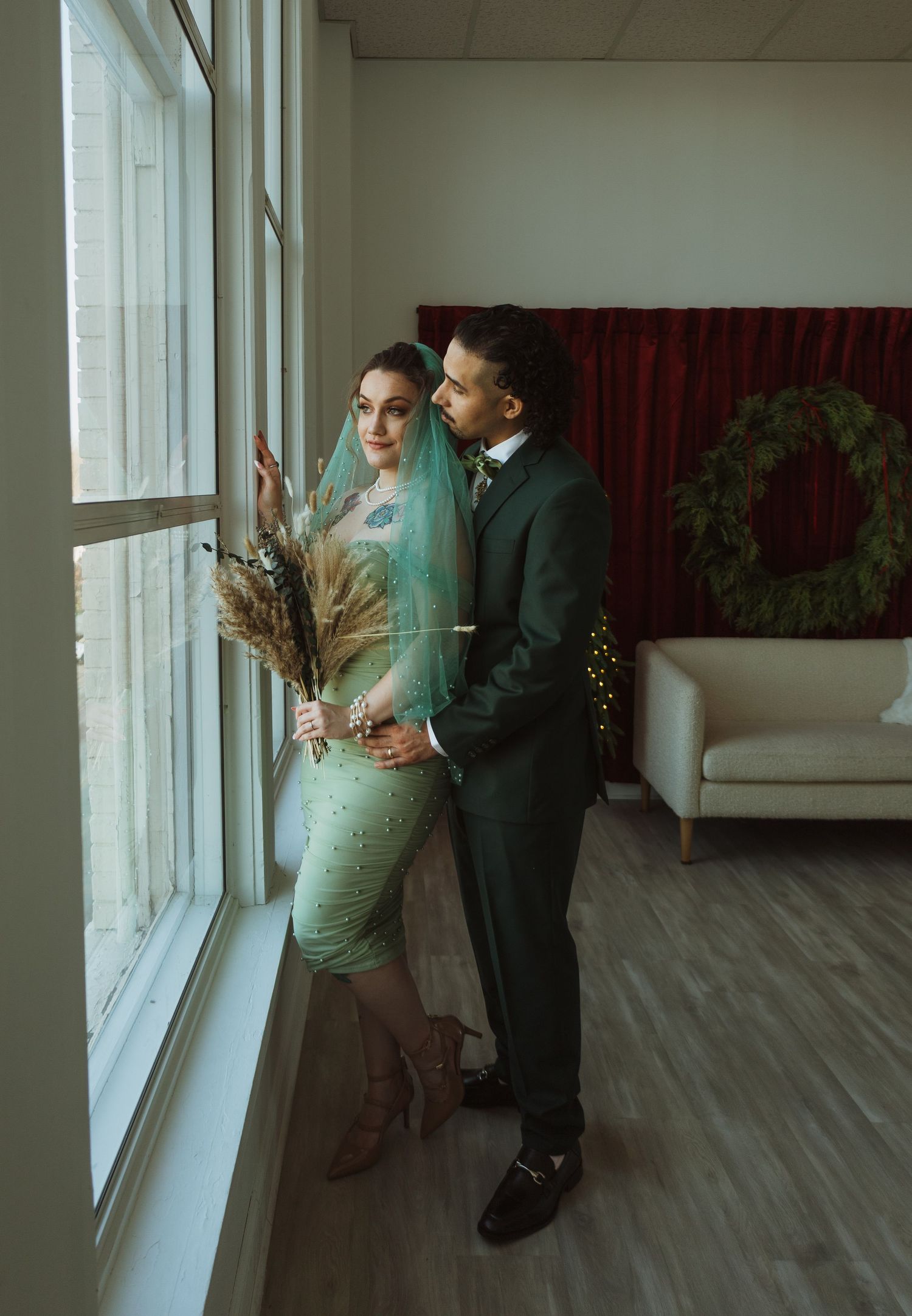 Bride and groom in coordinating green outfits gazing out window holding dried floral bouquet.