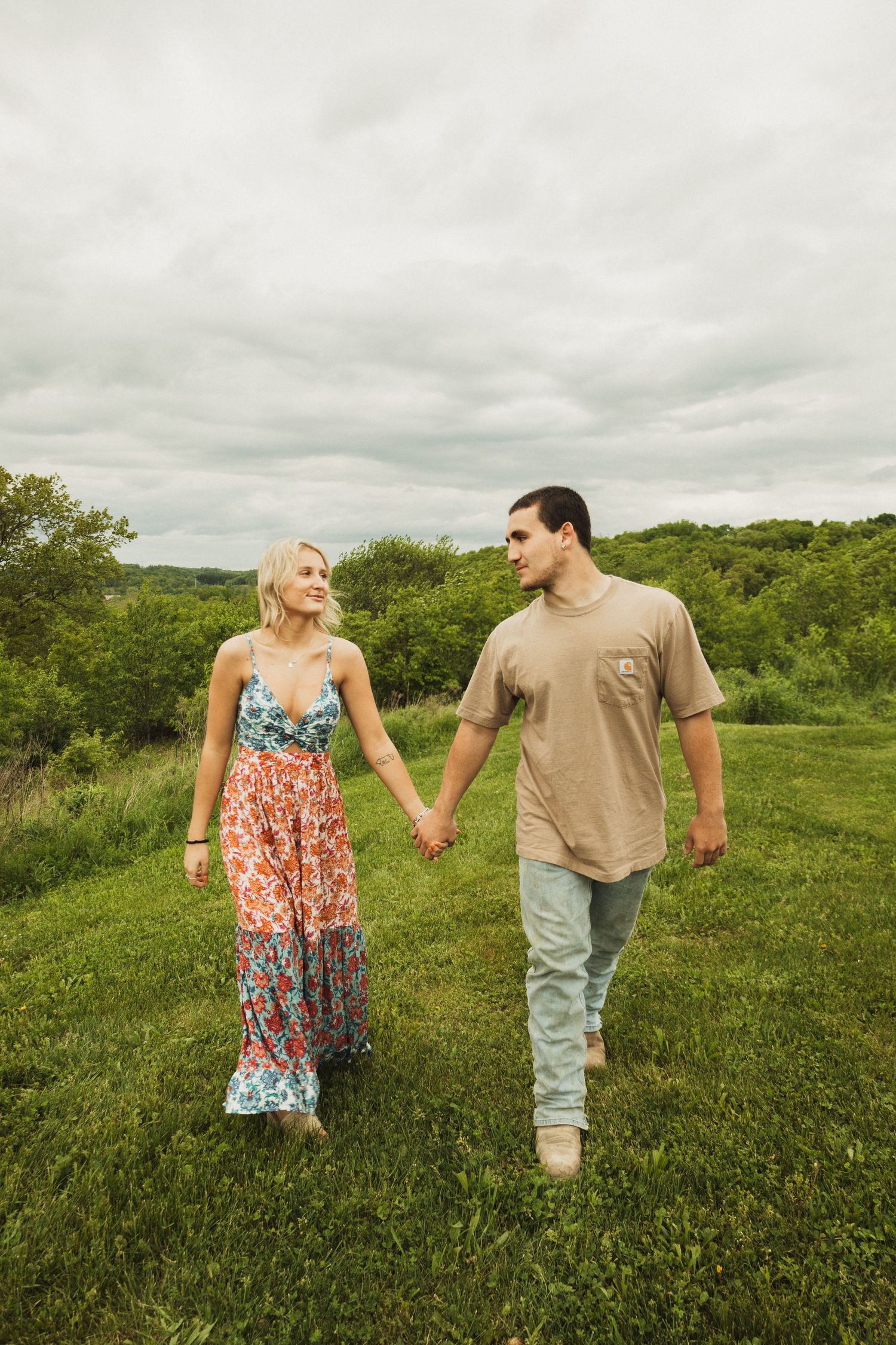 A couple holding hands while taking a romantic walk through a green hillside under cloudy skies.