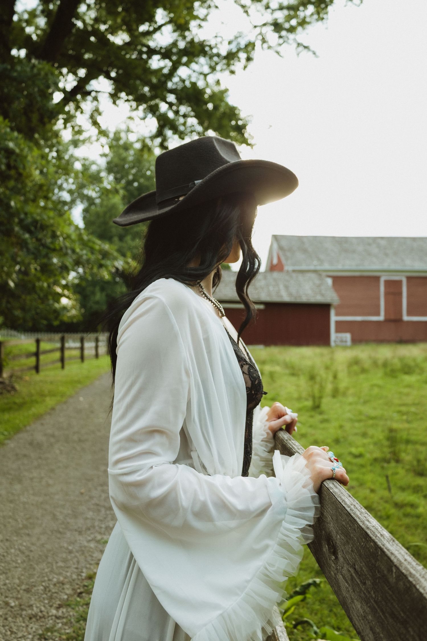 Someone in a white top and wide-brimmed hat leans against a wooden fence on a rural farm property.