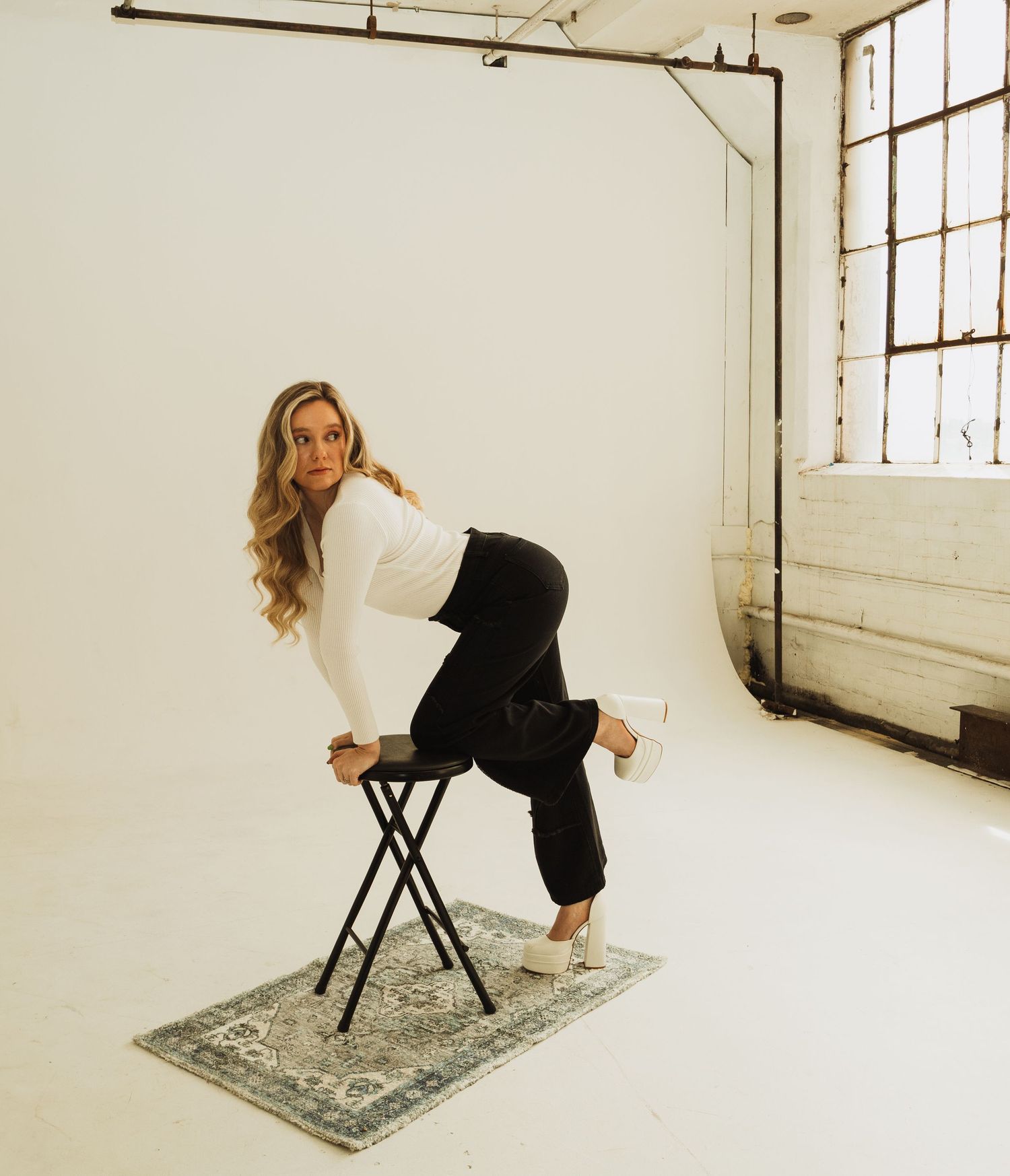 A model poses playfully on a black stool against a white studio backdrop with a decorative rug underneath.