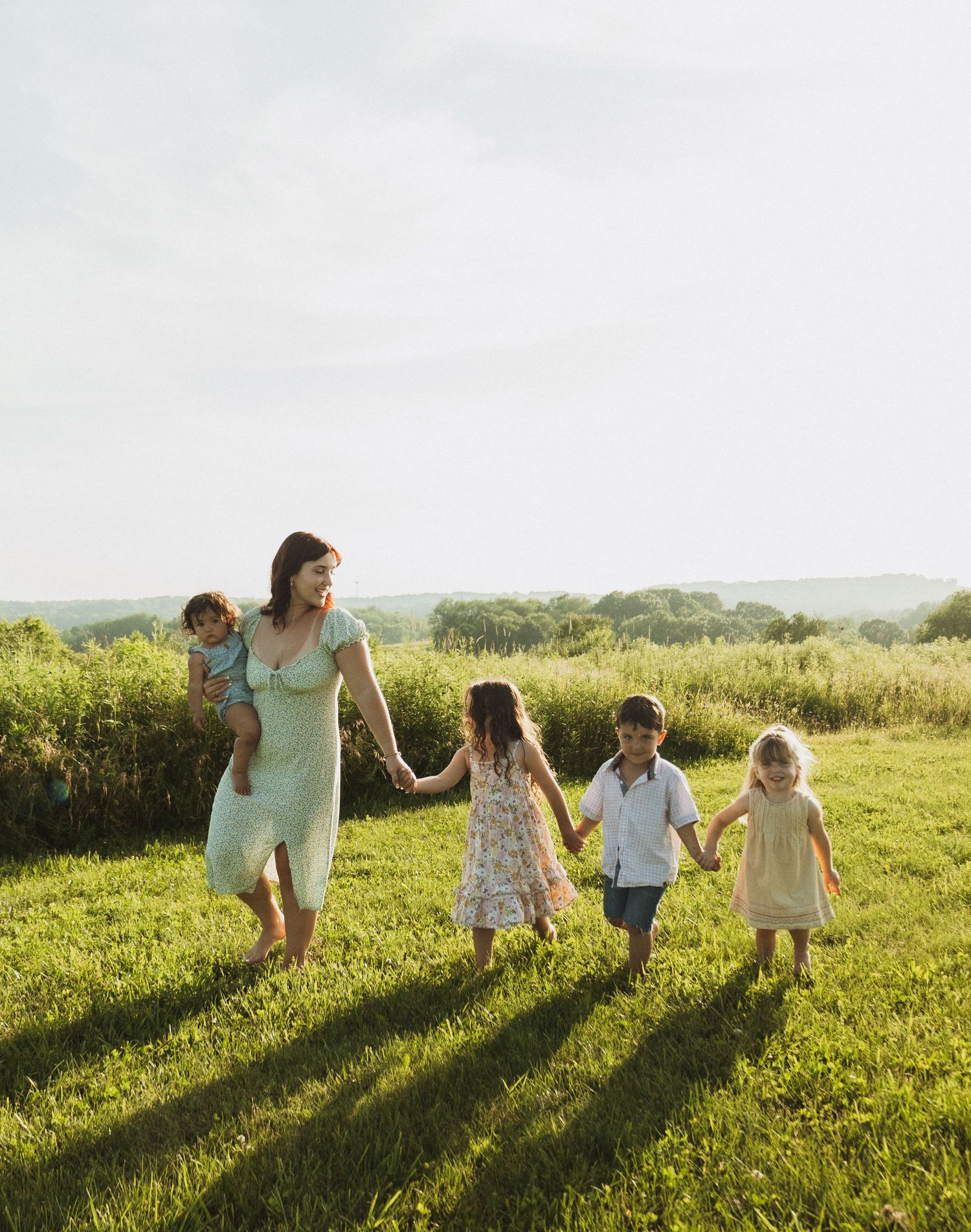 Group holds hands while walking through a green summer field at golden hour.